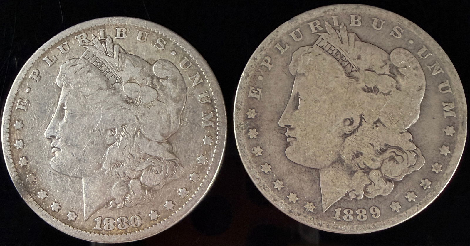 Two Morgan Silver Dollars, 1880 and 1889-O: Two United States Morgan silver dollars featuring examples dated 1880 and 1889. The obverse of each coin depicts a profile portrait of Liberty wearing a cap and crown inscribed with LIBERTY,