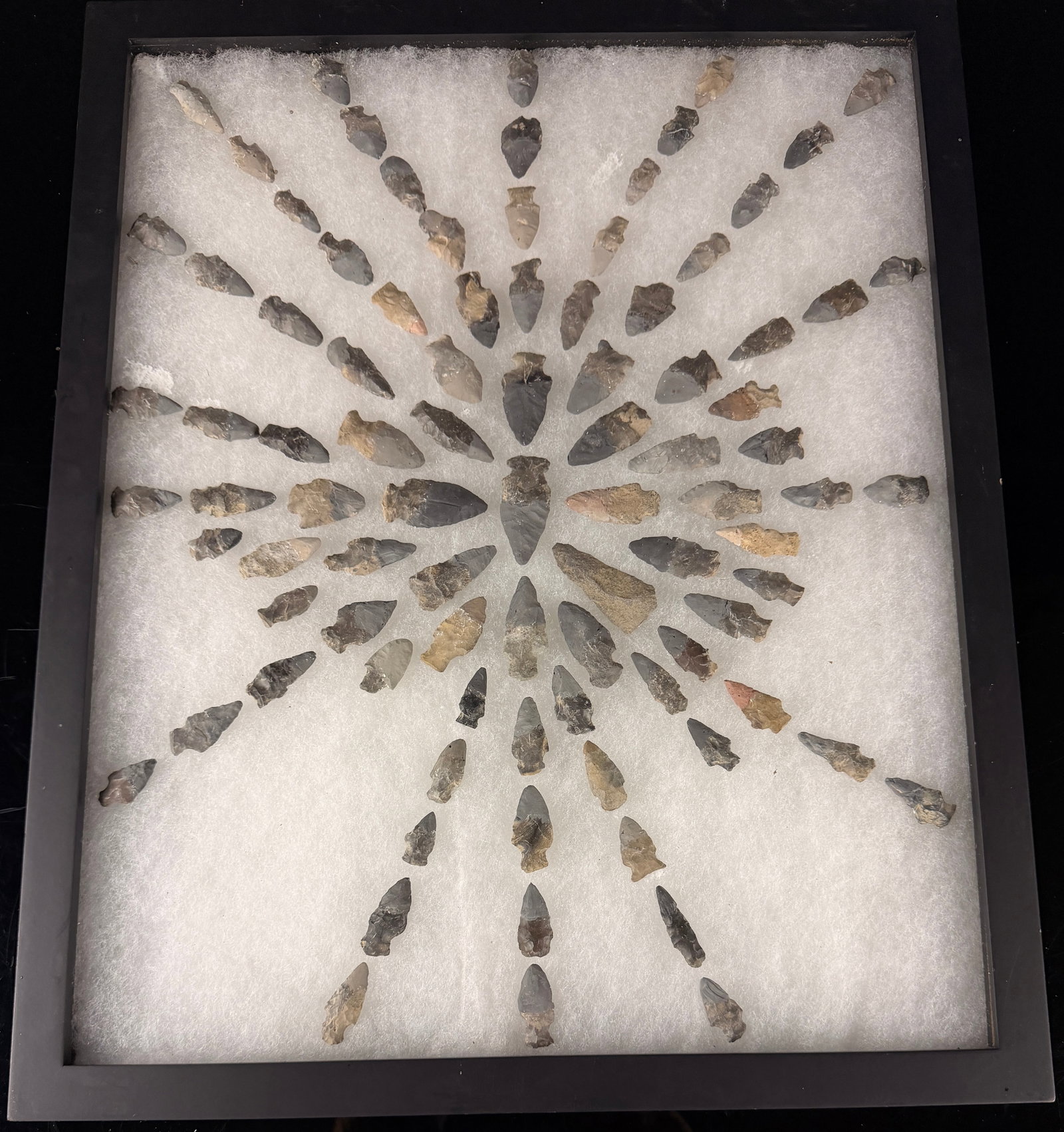 A Collection of Knapped Stone Lithic Points: This collection features a variety of knapped stone lithic points, commonly identified as arrowheads, arranged in a radiating starburst pattern. The points represent various typologies, including notc