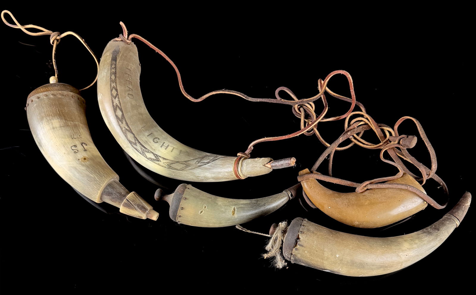 A Collection of American Carved Bovine Powder Horns: A collection of American hand-carved bovine black powder horns designed for use with muzzle-loading firearms. The items are constructed from translucent and opaque natural horn, featuring wooden end p