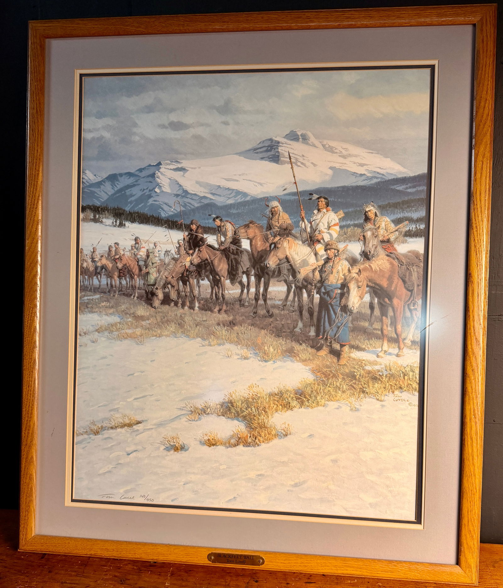 Tom Lovell The Blackfeet Wall Signed and Numbered Limited Edition Print: Tom Lovell, The Blackfeet Wall. This limited edition print on paper depicts a group of Native Americans on horseback traversing a snow-covered landscape with a large mountain peak in the background.
