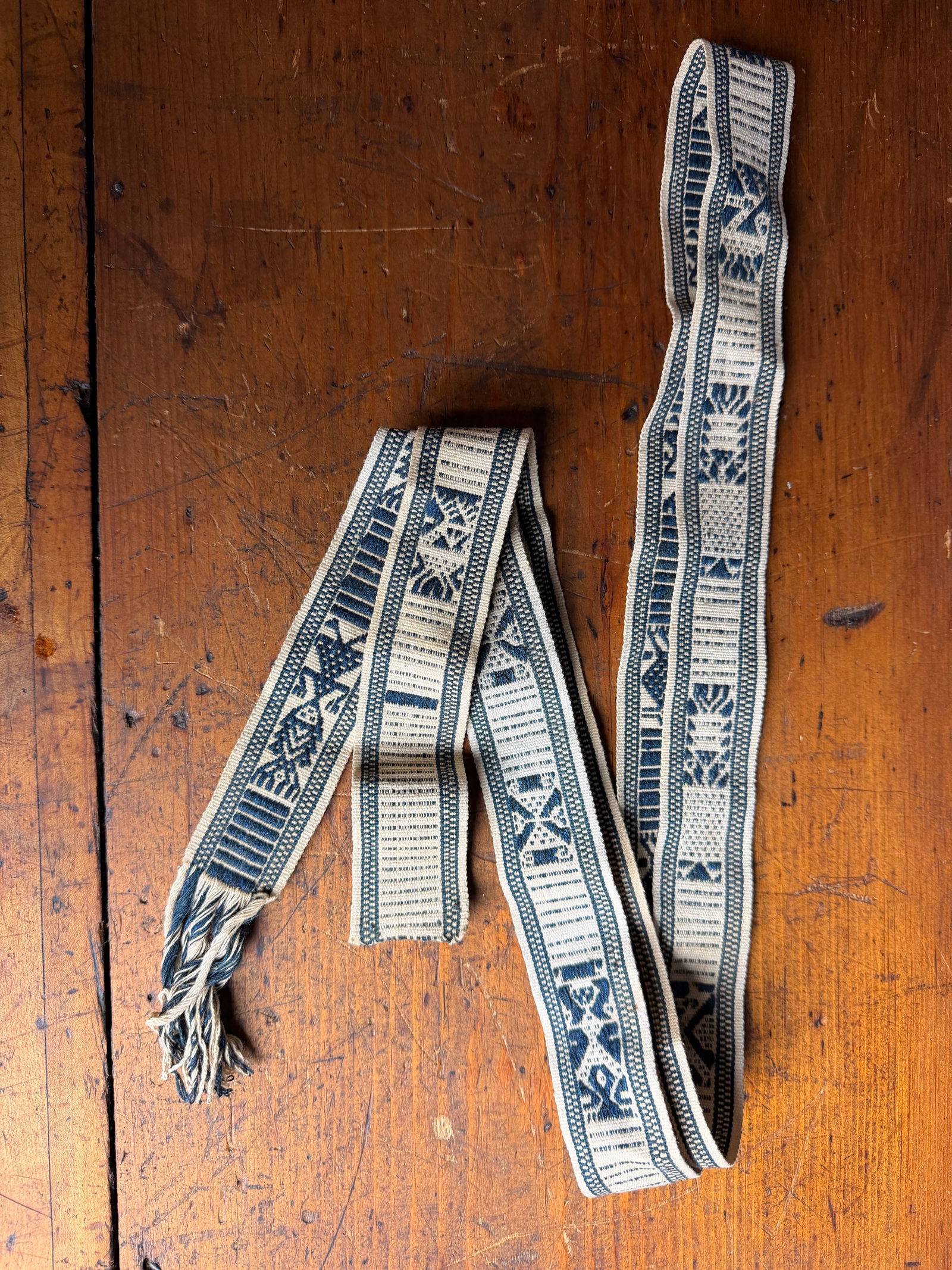 A Woven Indigo and Cream She People Textile Sash: This long, narrow textile strap is a traditional woven sash attributed to the She people (Shezu) of China. The piece is constructed using a decorative weave in cream and indigo blue. The design