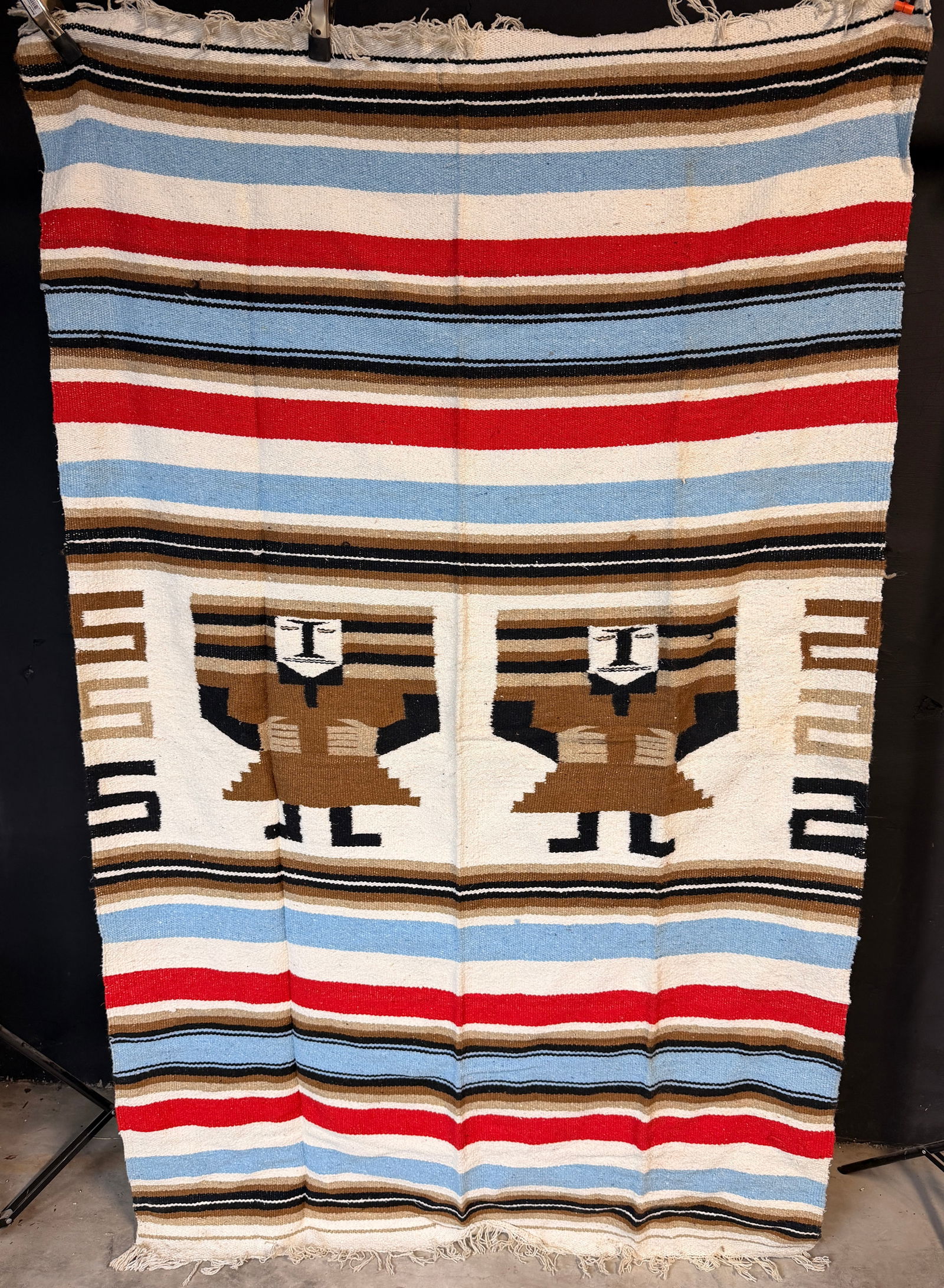 A Vintage Mexican Wool Pictorial Rug: A Mexican woven wool pictorial rug or wall hanging. The textile features a central cream field decorated with two stylized human figures wearing brown and black garments, flanked by geometric stepped