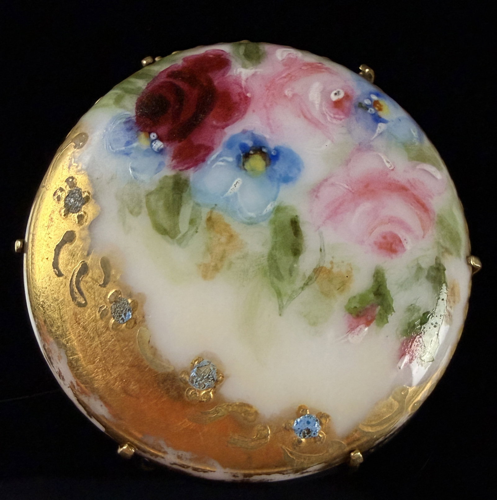 Hand-Painted Porcelain Floral Brooch with Gilded Scrollwork: This circular brooch features a hand-painted porcelain cabochon in the style of Limoges, depicting a floral bouquet of crimson and pink roses, blue blossoms, and green foliage against a white ground.