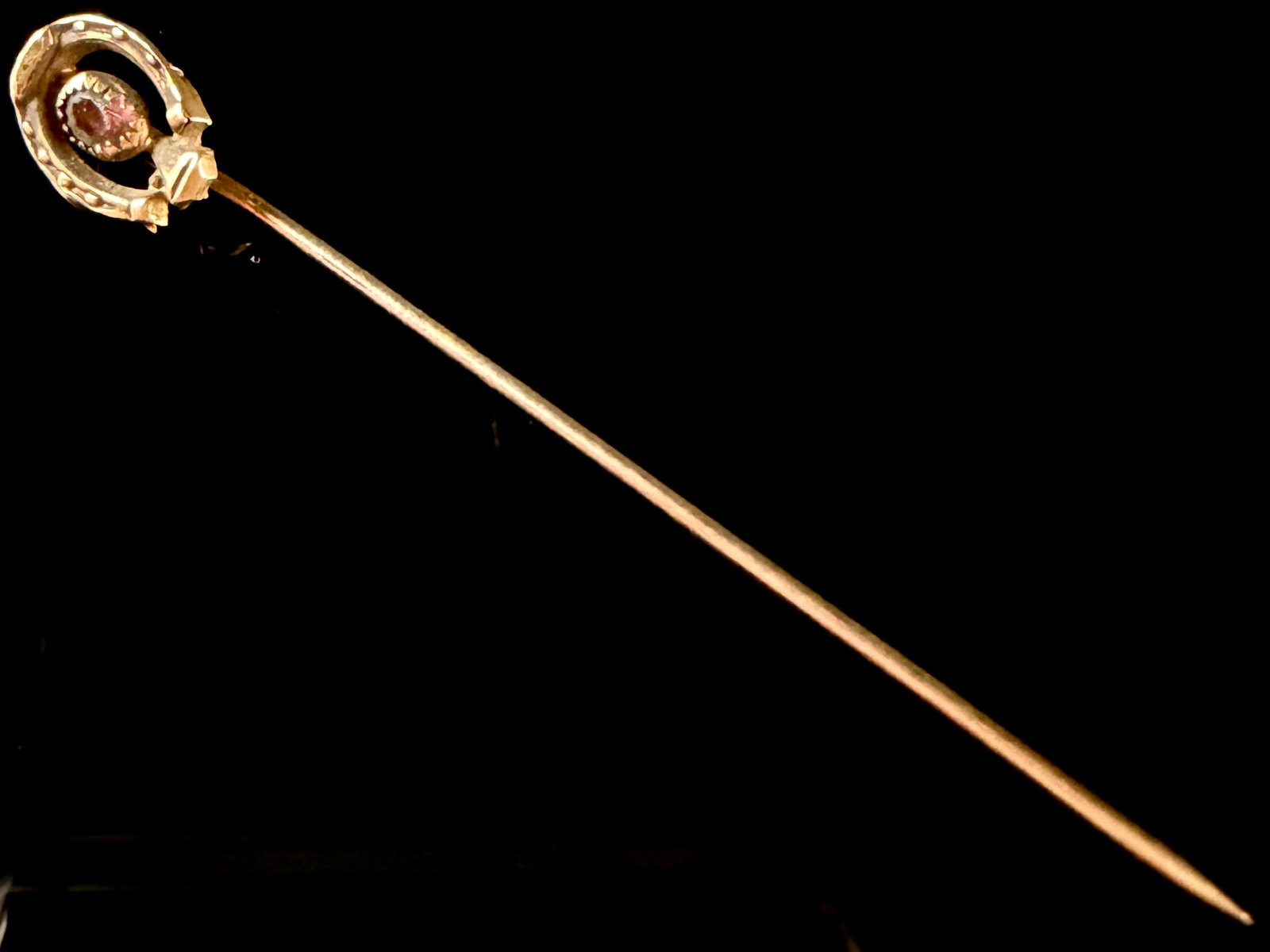 Victorian Style Horseshoe Stickpin with Purple-Red Gemstone: This stickpin features an equestrian horseshoe motif crafted from yellow gold-tone metal. Centered within the horseshoe is a single oval faceted purple-red gemstone, resembling a garnet or amethyst, s