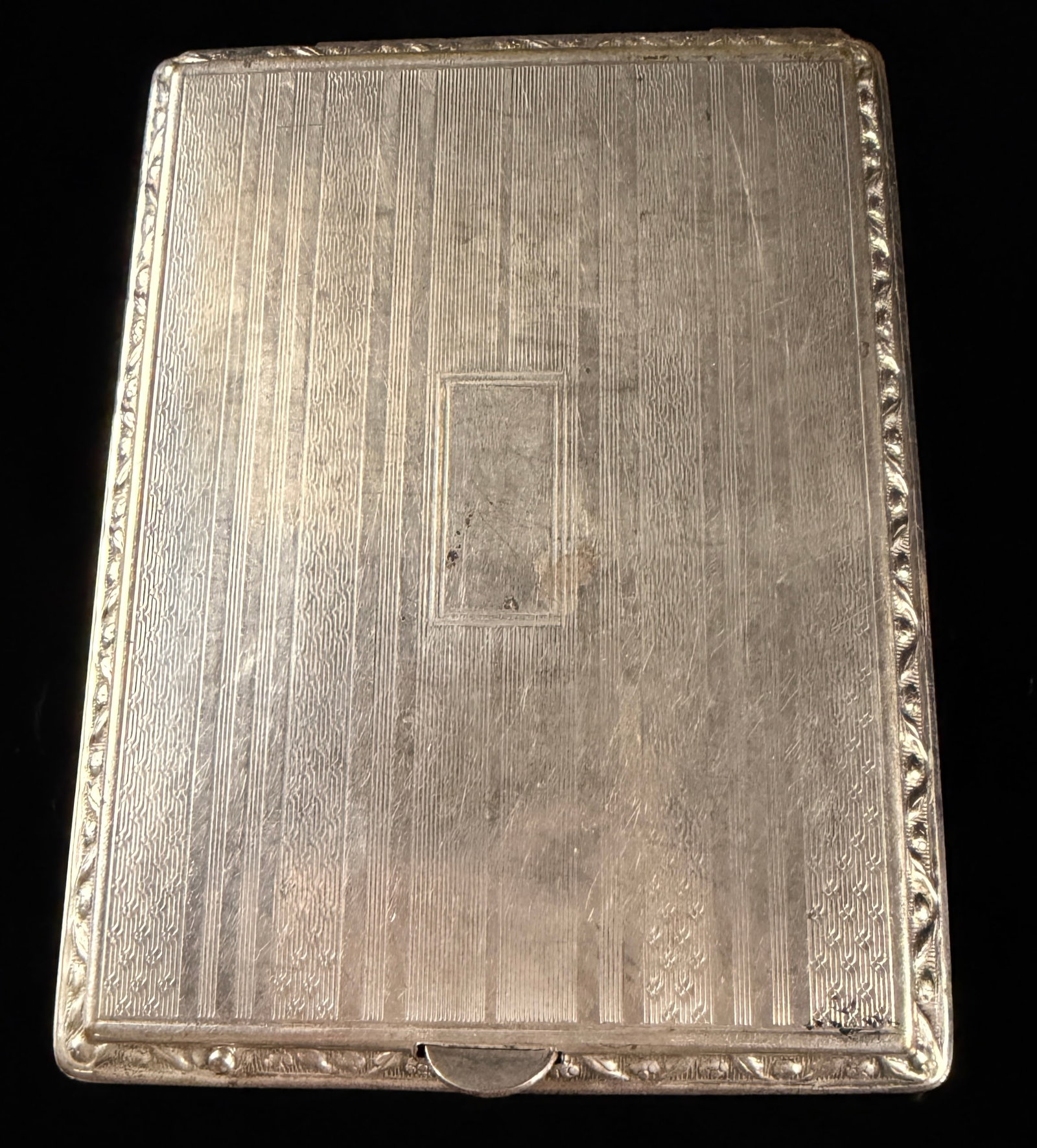 Art Deco Silver-Toned Engine-Turned Cigarette Case with Foliate Border: This rectangular hinged cigarette case is designed in the Art Deco style. The exterior displays an engine-turned guilloche pattern consisting of alternating vertical bands of fine parallel lines and t