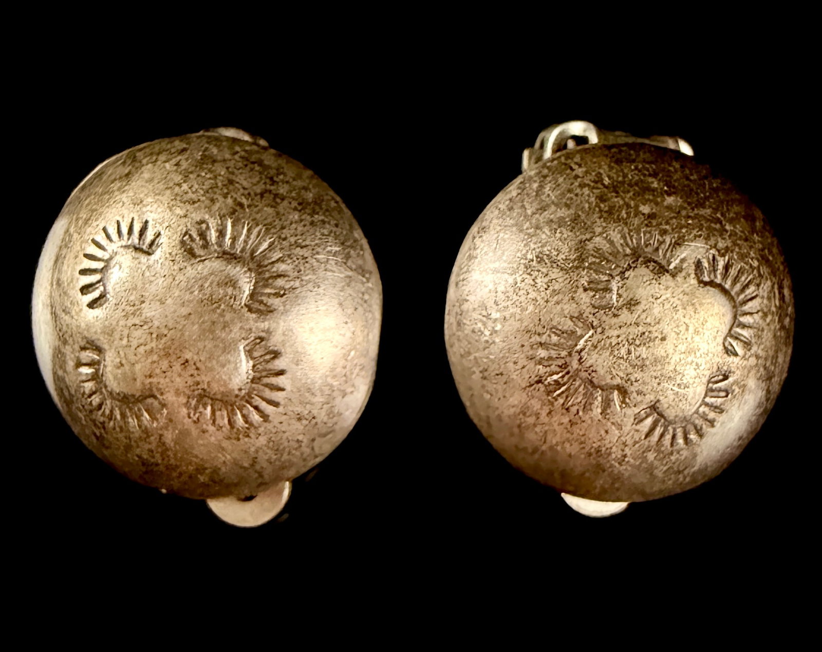 A Pair of Southwestern Style Stamped Silver Tone Domed Clip On Earrings: A matched pair of clip-on earrings designed in a domed circular button style. The earrings are made of silver-toned metal and feature stamped motifs resembling sunbursts or crescents on the front surf
