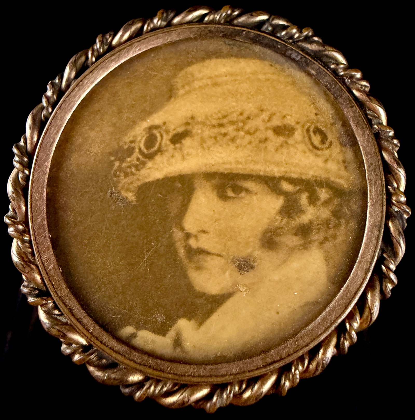 Antique Gold-Toned Circular Photographic Portrait Brooch with Twisted Rope Border: This circular brooch features a sepia-toned photographic portrait of a young woman shown in profile. The subject is depicted wearing a wide-brimmed hat with floral or lace appliqu?©s, characteristic