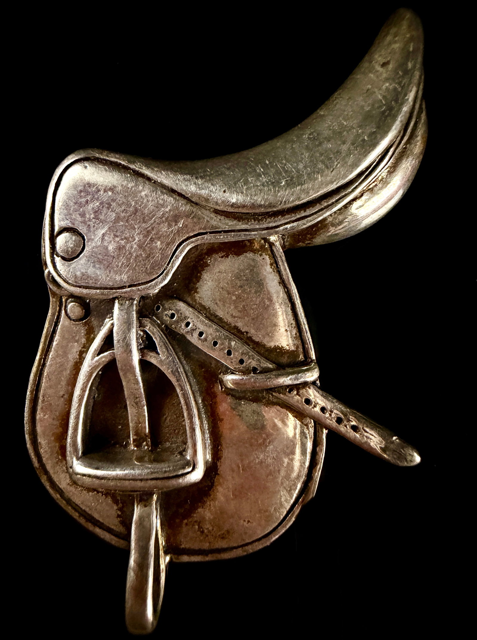 Sterling Silver English Riding Saddle Brooch: A sterling silver brooch cast in the form of an English-style riding saddle. The piece features realistic details including a hanging stirrup and a girth strap with punched holes. The reverse is fitte