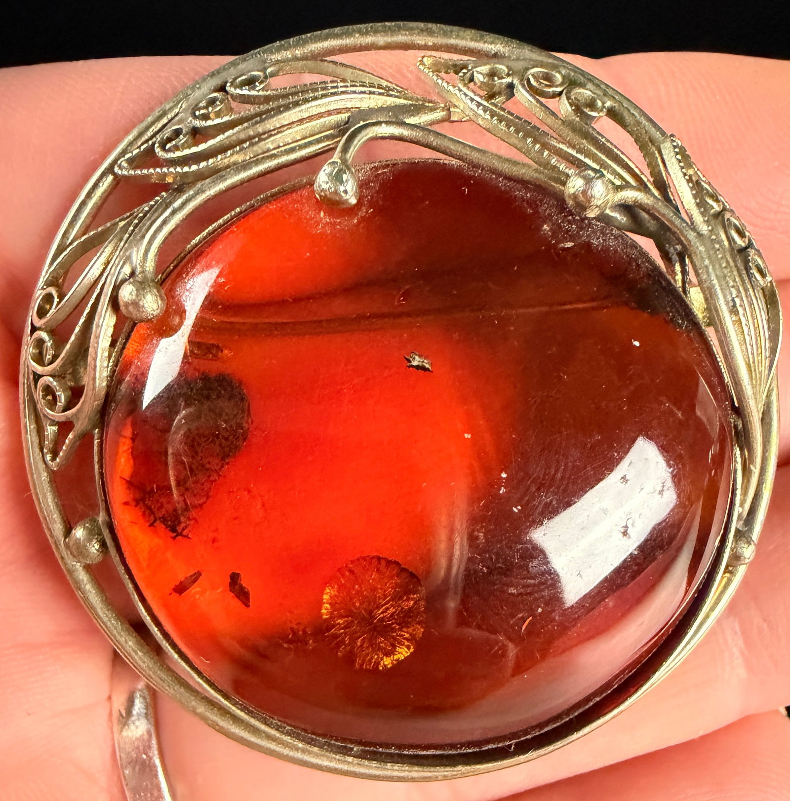 European .835 Silver and Baltic Amber Circular Openwork Brooch: A circular openwork brooch featuring a large central cabochon of Baltic amber. The amber is a deep reddish-orange color and contains internal circular stress fractures known as sun spangles. The stone