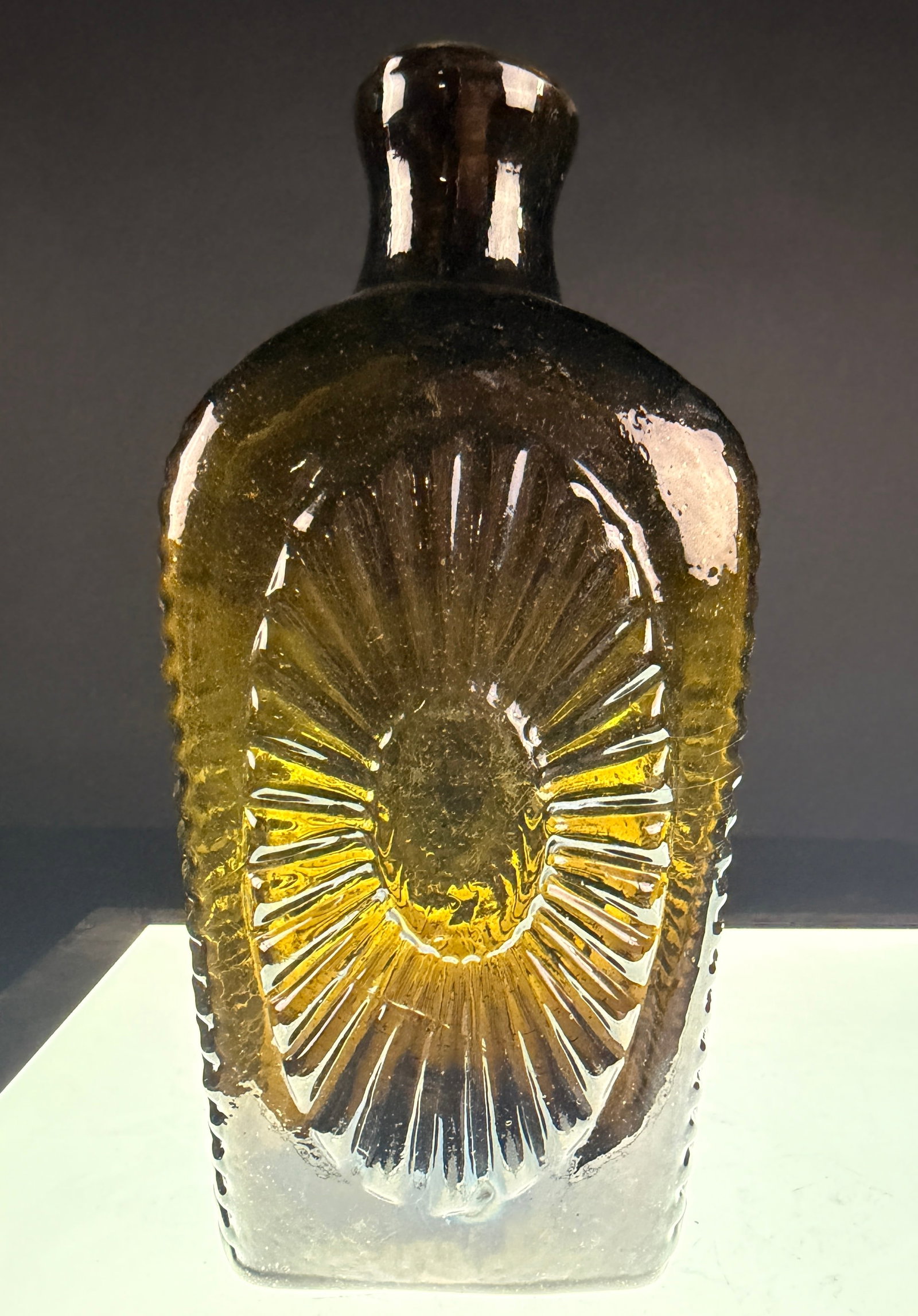 Early American Olive Amber Mold Blown Sunburst Glass Flask: Olive-amber mold-blown glass flask featuring ribbed sides, a central sunburst motif on both faces, and a rough pontil mark, measuring approximately 5.5 inches in height.