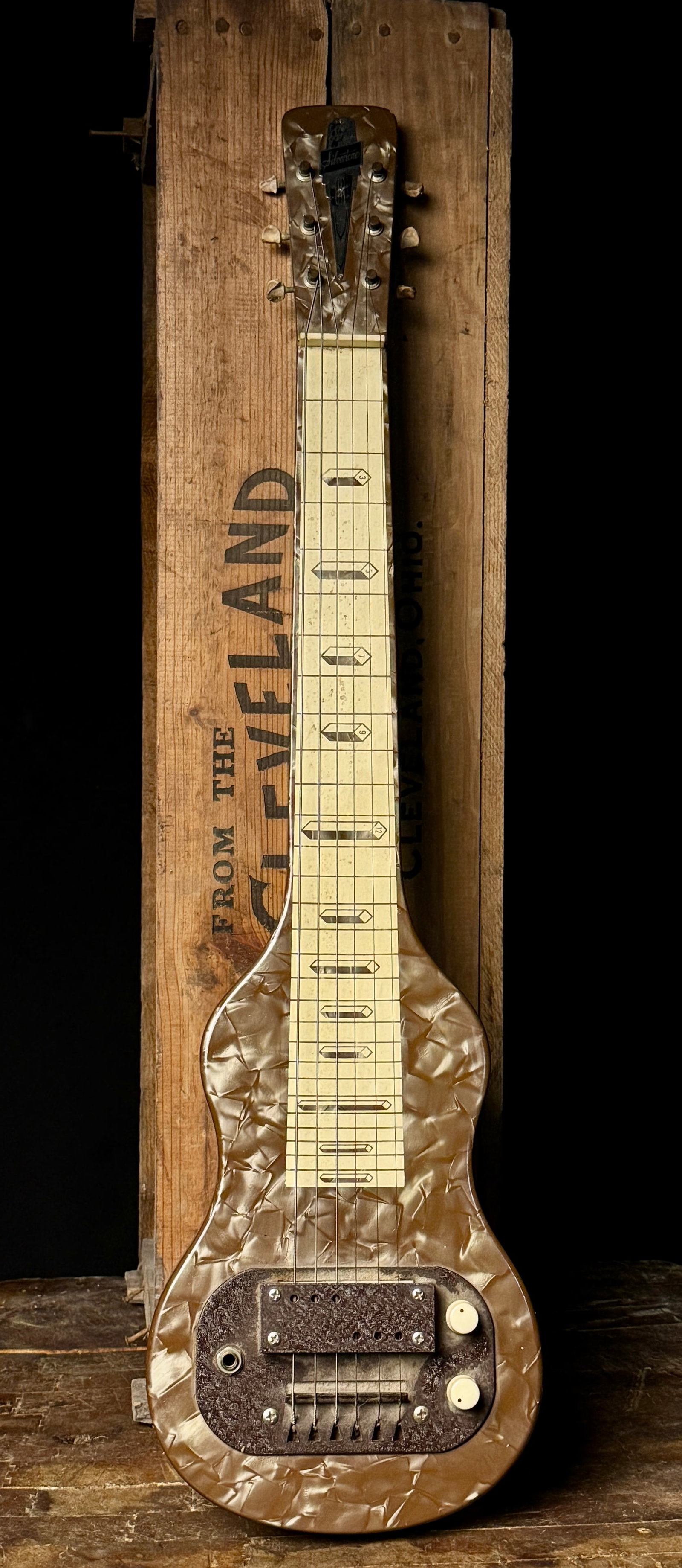 Silvertone Pearloid Lap Steel Guitar Manufactured by Valco Serial X52199: This vintage six-string lap steel guitar features the Silvertone brand name on a decorative Art Deco metal headstock plate. The instrument is constructed with a wooden body and neck wrapped in a brown
