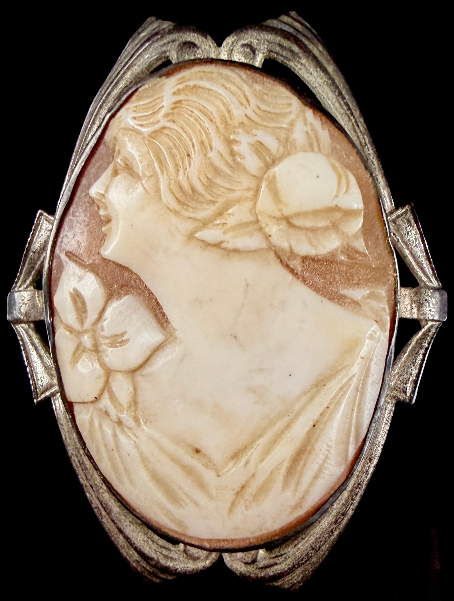 Art Deco Silver Tone Shell Cameo Brooch with Openwork Frame: An Art Deco period jewelry piece featuring a hand-carved shell cameo. The oval cameo depicts a classical female profile in relief, facing left, with a prominent floral motif at the shoulder and hair s