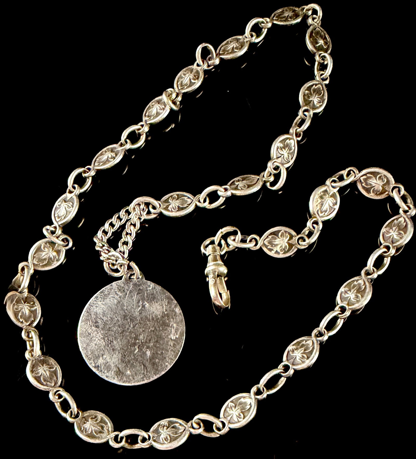 Silver-Toned Foliate Link Necklace with Swivel Clasp and Circular Pendant: A silver-toned necklace featuring fancy oval links decorated with a raised three-lobed foliate motif. The chain is secured with a traditional swivel-style dog clip clasp. A plain, flat, circular penda