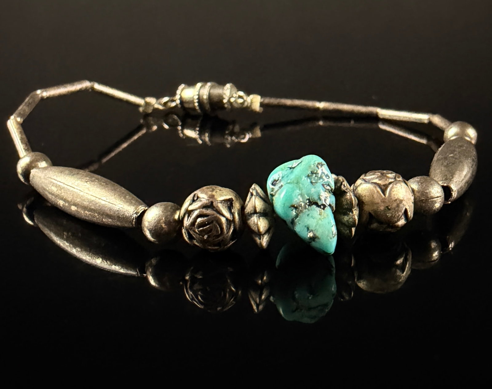 Southwestern Style Silver-Tone and Turquoise Nugget Beaded Bracelet: A beaded strand bracelet featuring a central irregularly shaped blue-green stone nugget resembling turquoise. This centerpiece is flanked by two ornate beads with rose motifs and two elongated, tapere