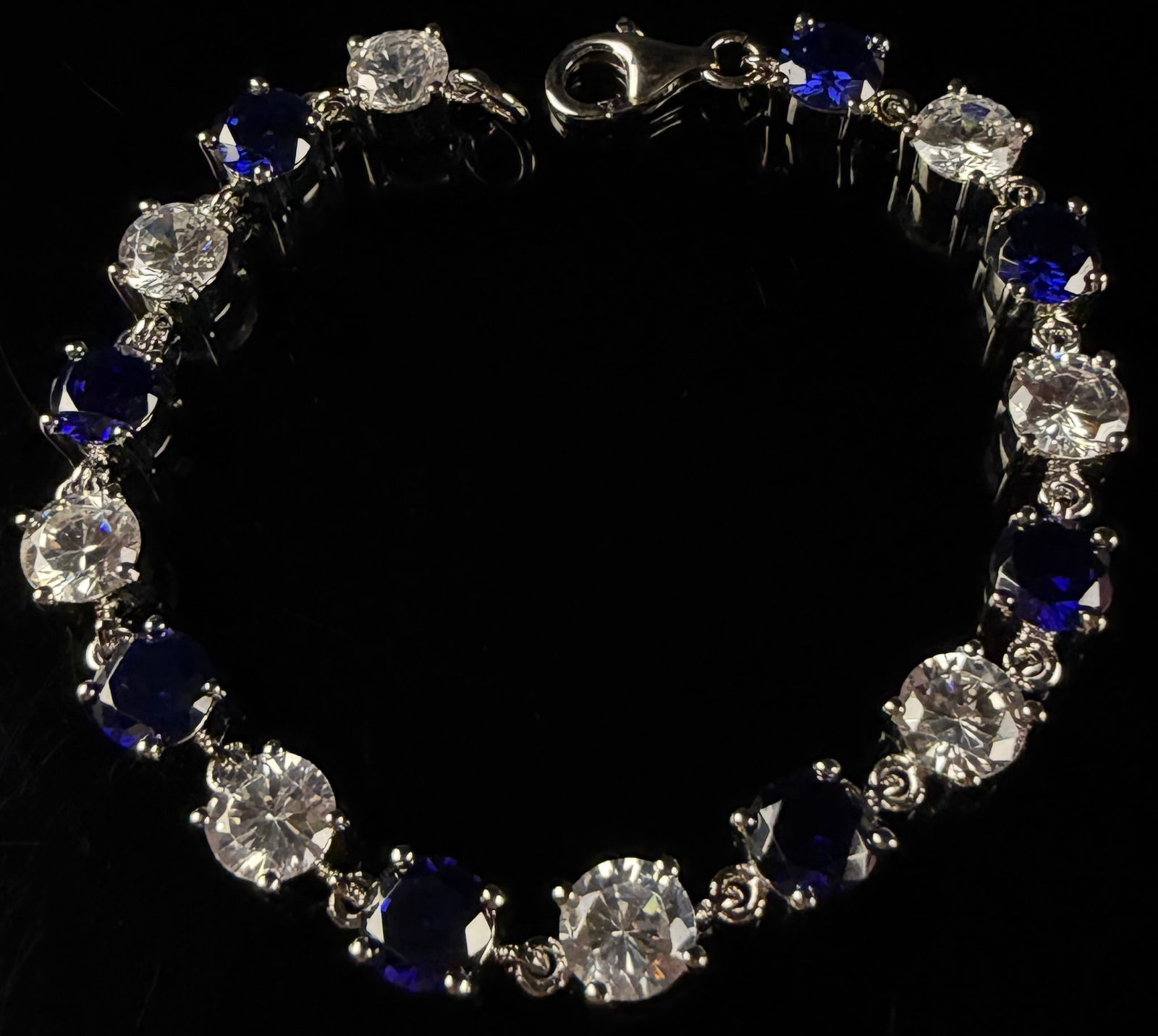 Sterling Silver Tennis Bracelet with Alternating Blue and Colorless Stones: This sterling silver tennis bracelet features seventeen circular articulated links in a classic linear design. The bracelet is set with an alternating pattern of nine deep blue round-cut faceted stone