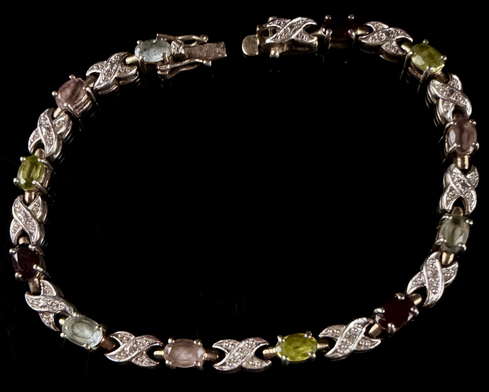 ICC Sterling Silver Multi-Gemstone and Accent Stone X-Link Tennis Bracelet: This multi-gemstone tennis bracelet is constructed of sterling silver with a repeating X-link design. The piece features a series of prong-set, oval-cut gemstones in a variety of colors including deep