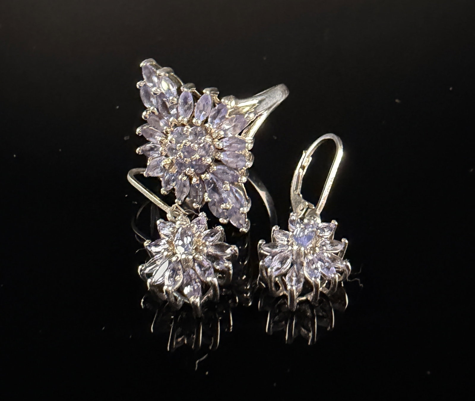 A Sterling Silver and Lavender Gemstone Floral Motif Jewelry Suite: A jewelry collection consisting of a statement ring and a matching pair of lever-back drop earrings, all constructed in sterling silver. Each piece features faceted lavender-colored gemstones in marqu