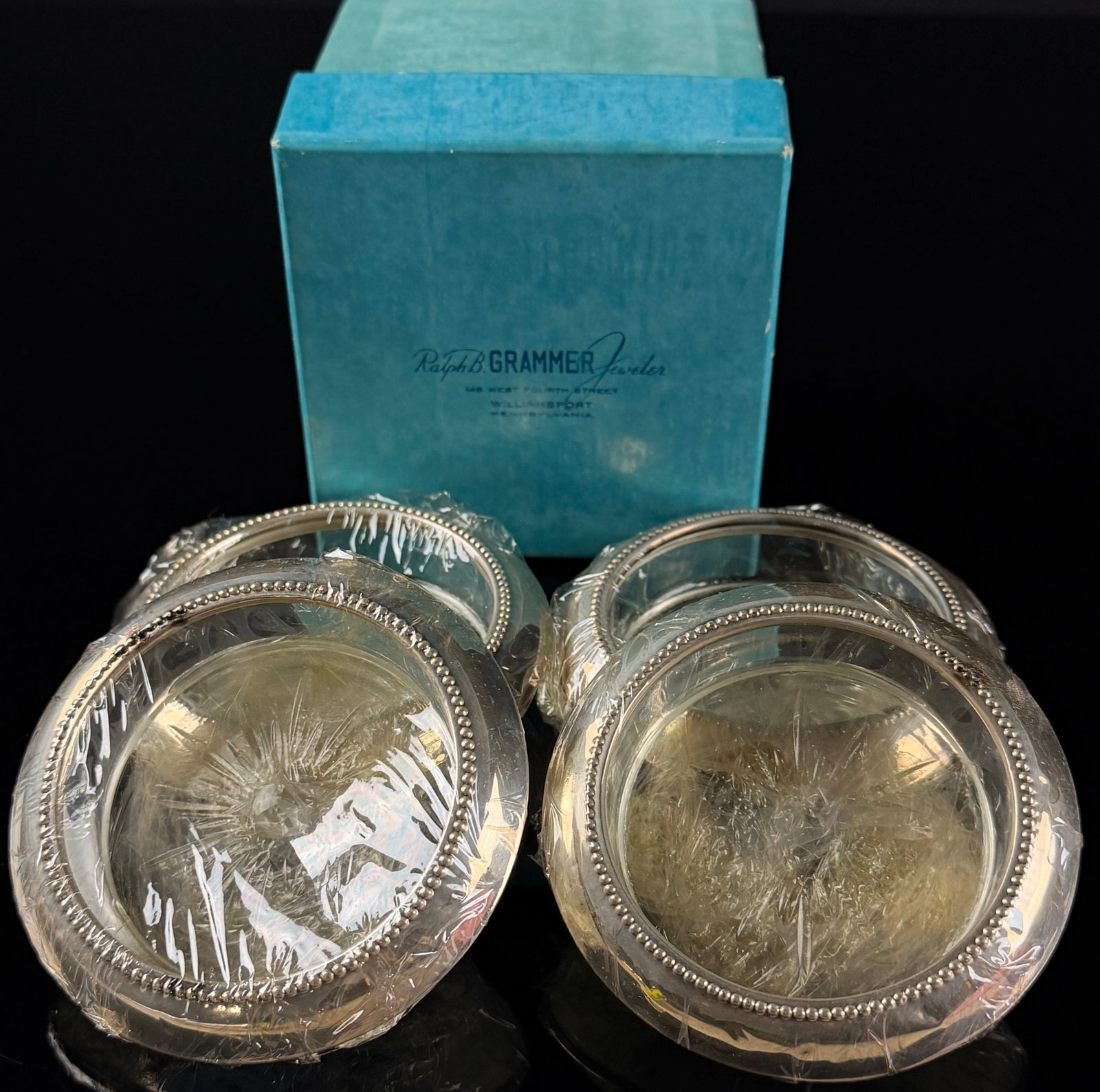 A Set of Four Sterling Silver and Glass Coasters: A set of four circular coasters featuring clear glass centers and sterling silver rims. Each coaster is designed with a recessed glass base that displays a molded starburst or sunburst pattern on the