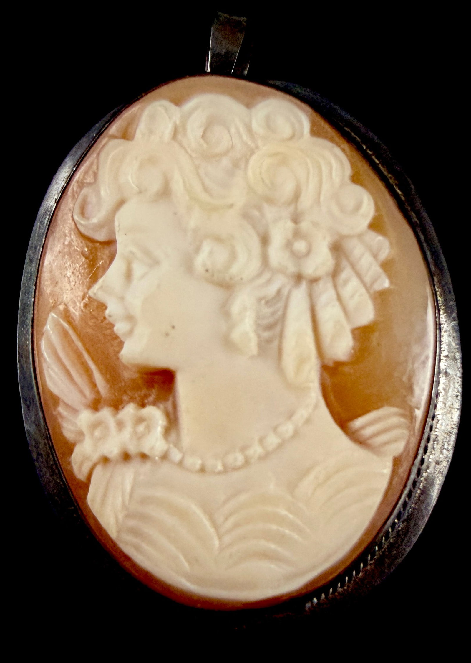 Italian Carved Shell Cameo Brooch-Pendant in Sterling Silver: An Italian hand-carved shell cameo brooch-pendant bezel-set in a sterling silver frame. The oval cameo depicts a left-facing profile of a young woman with elaborately styled hair featuring floral acce
