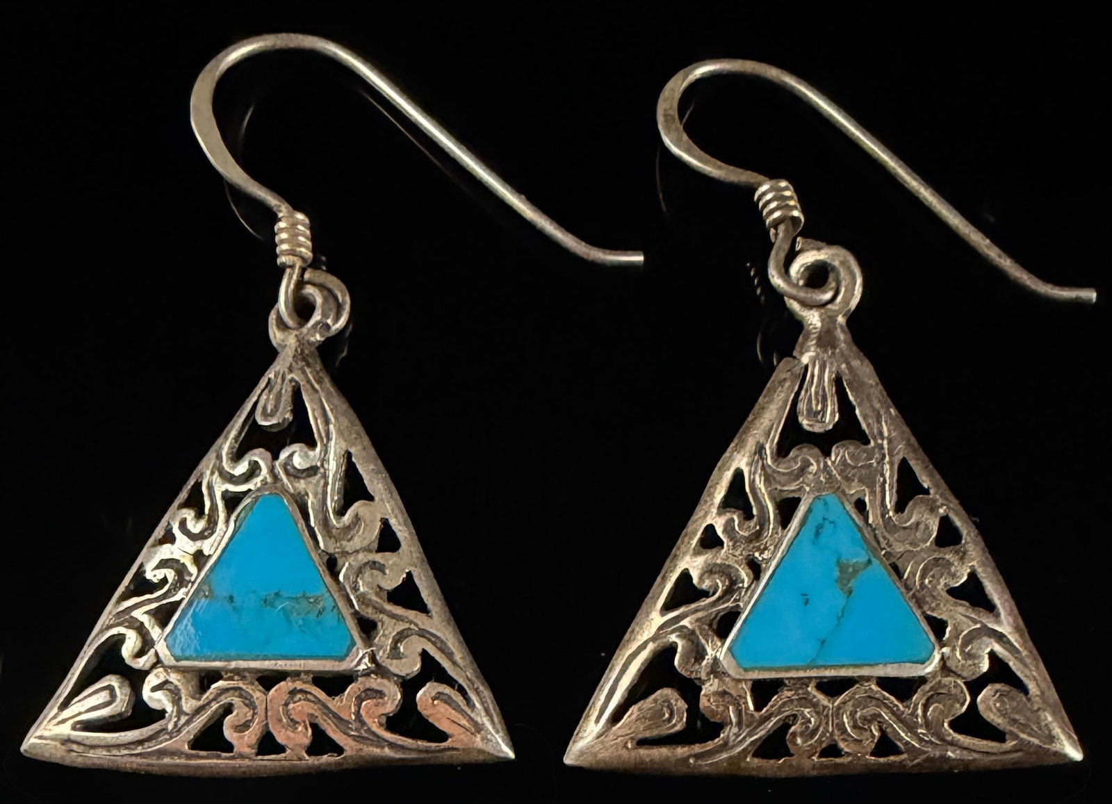 A Pair of Sterling Silver and Turquoise Triangular Filigree Drop Earrings: A matched pair of sterling silver drop earrings featuring a triangular openwork design. The silver pendants are decorated with intricate scroll and filigree metalwork. Each earring contains a central 