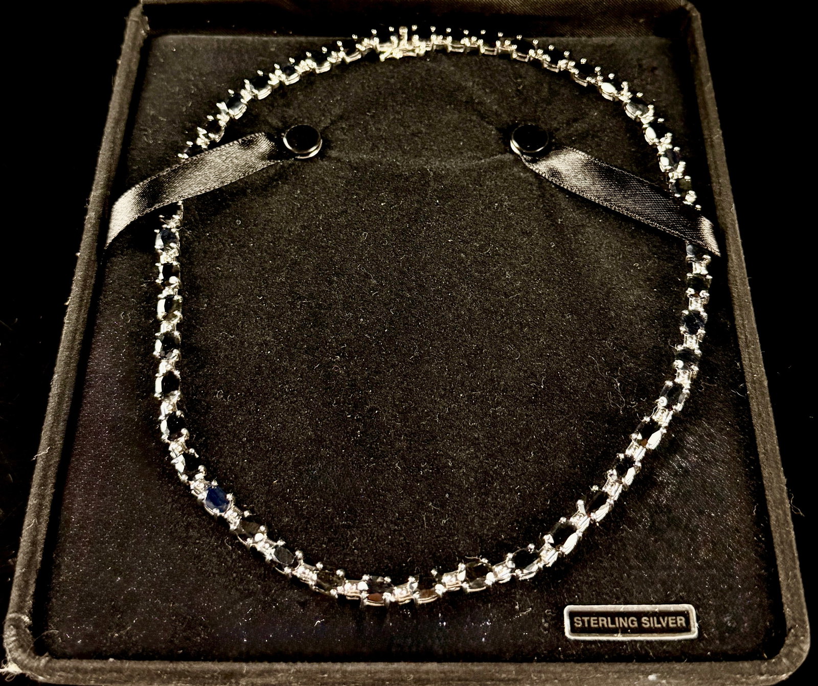 G.L.O. Sterling Silver and Blue Gemstone Tennis Necklace: This is a sterling silver tennis-style necklace manufactured by G.L.O. Jewelry. The design features a continuous line of oval-cut dark blue gemstones alternating with vertical baguette-cut clear