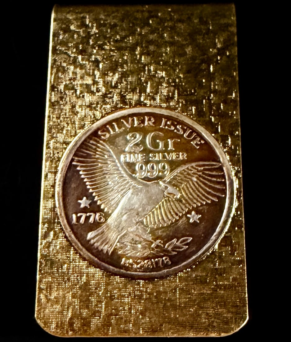 US Bicentennial 2 Gram .999 Fine Silver Round Gold Tone Money Clip: This gold-tone metal money clip features an inset .999 fine silver round produced as a souvenir for the 1976 United States Bicentennial. The clip features a textured, cross-hatched surface pattern on 