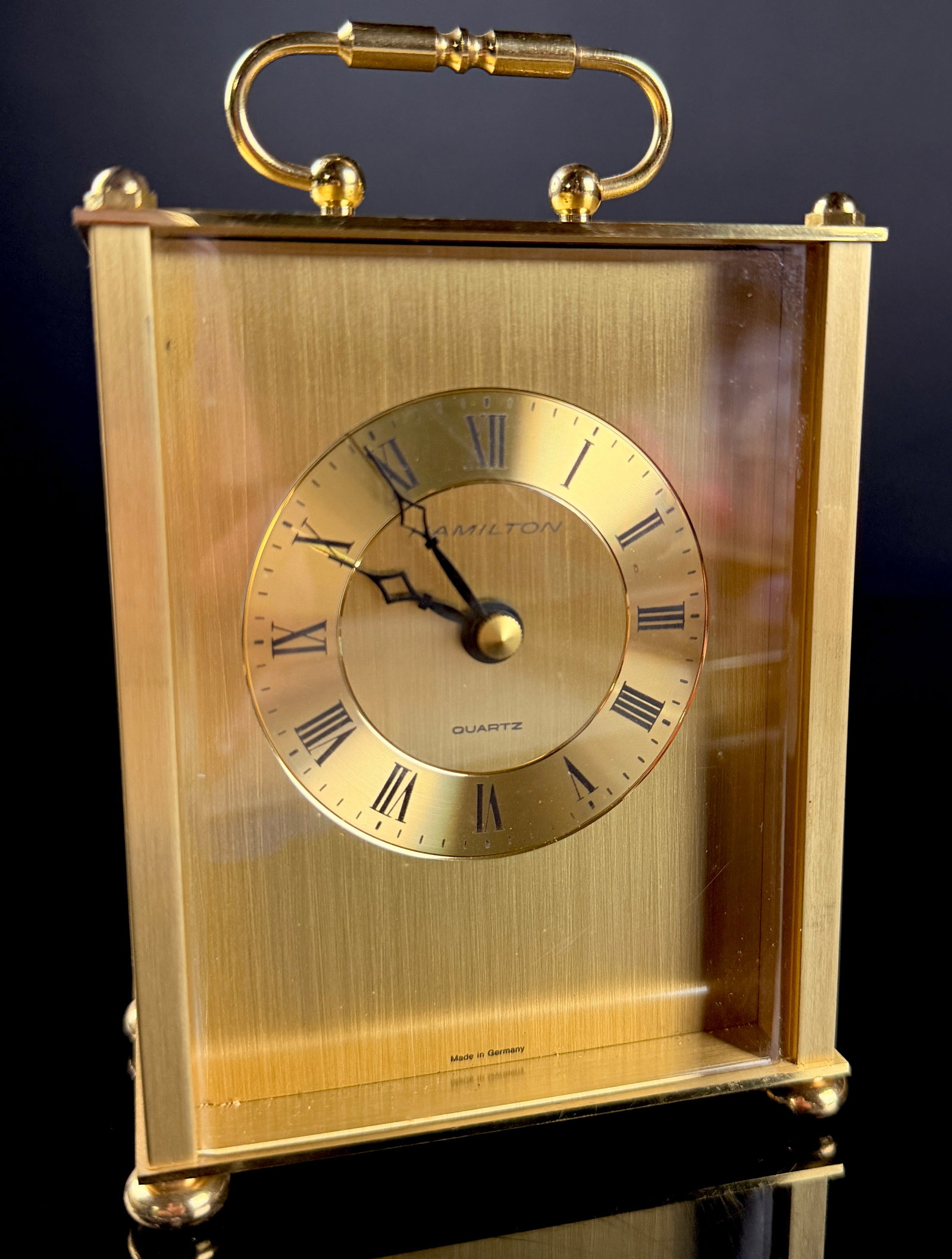 Hamilton Brass Quartz Carriage Clock West Germany: A brass carriage clock manufactured by Hamilton. The piece features a gold-toned rectangular case with a vertical brushed finish, a hinged rear door, a top swing handle, and four bun feet. Materials f