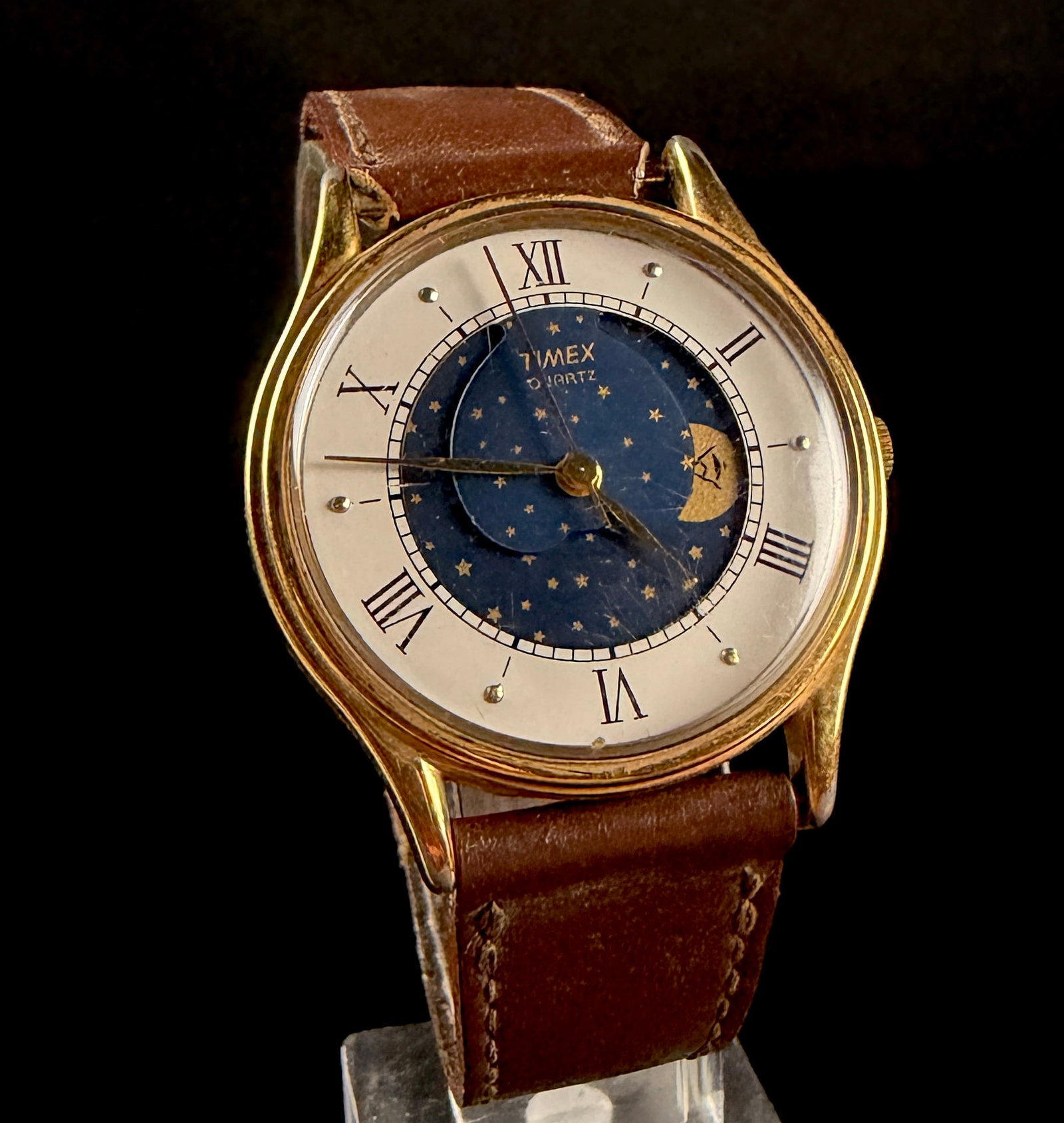 Vintage Timex Gold Tone Quartz Moonphase Wristwatch: A vintage Timex quartz wristwatch featuring a day and night celestial complication. The watch is constructed with a gold-tone base metal case. The two-tone dial consists of a white outer ring with bla