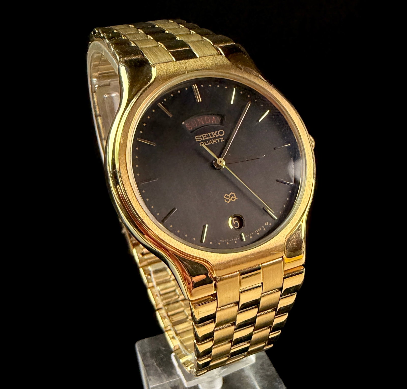 Seiko Quartz Day-Date Dress Watch 5Y23-7159: A Seiko quartz dress watch, reference number 5Y23-7159. The timepiece features a round gold-toned base metal case with a matching integrated multi-link bracelet. The black dial is detailed with applie