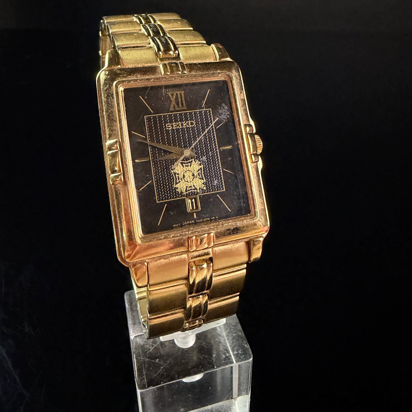 Seiko Veterans of Foreign Wars Glen M Gardner Jr Commemorative Gold Tone Quartz Wristwatch: A Seiko quartz wristwatch featuring a rectangular gold tone stainless steel case and integrated link bracelet. The black dial is designed with a textured center panel featuring a gold tone Veterans