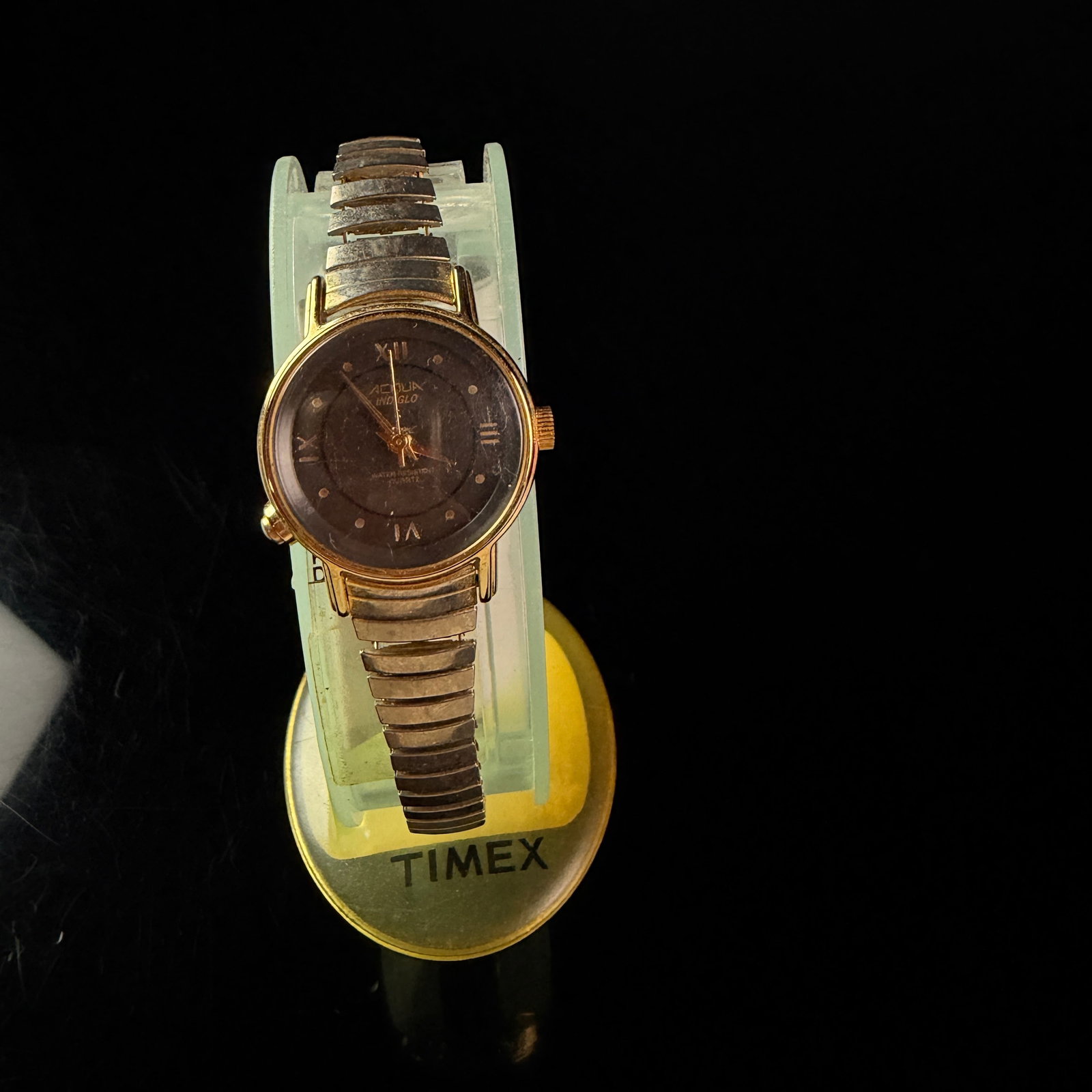 Ladies Acqua by Timex Indiglo Quartz Wristwatch with Expansion Band: This is a ladies quartz wristwatch produced by Acqua, a sub-brand of Timex. The timepiece features a round gold-tone base metal bezel and a stainless steel case back. The dark brown dial is designed