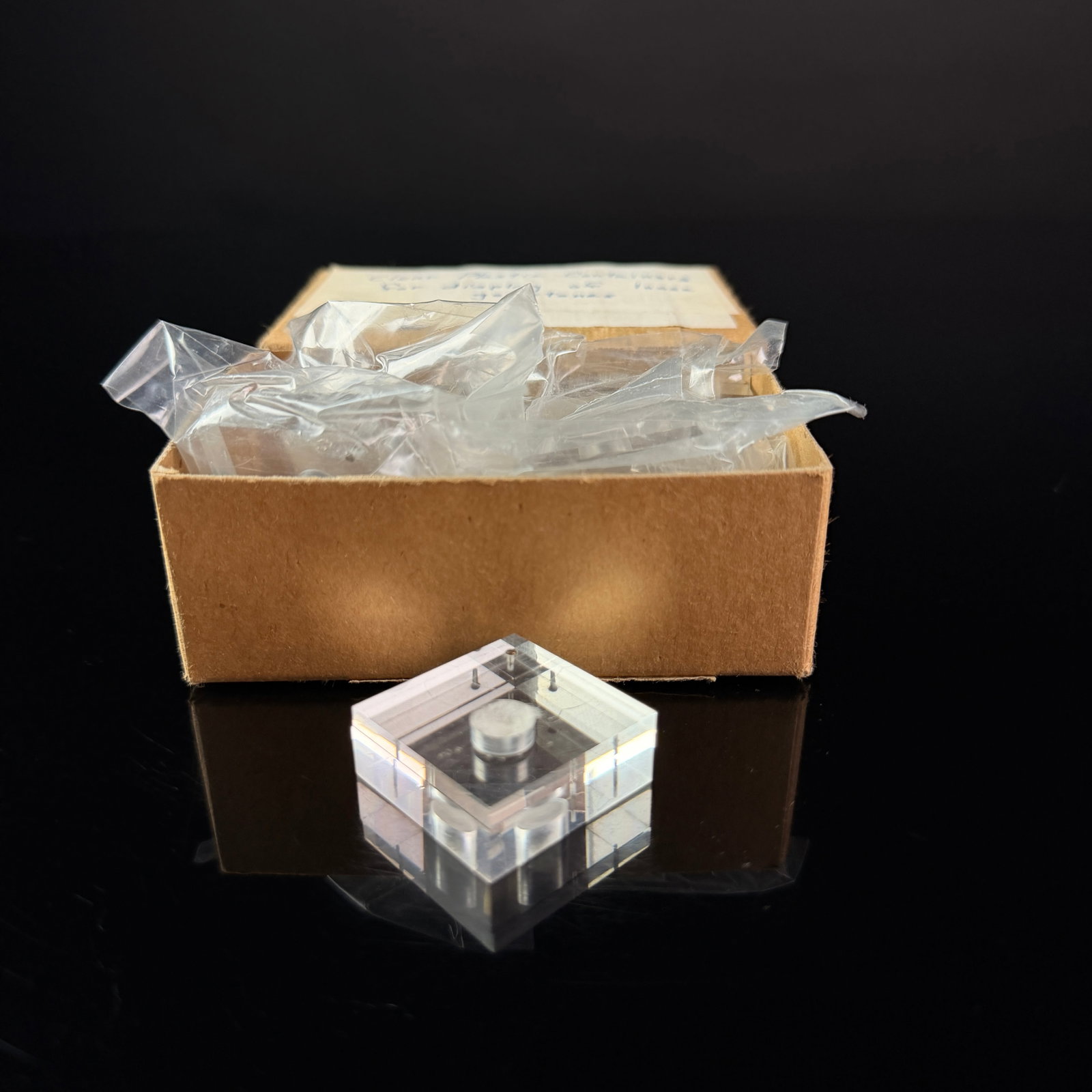 A Collection of Vintage Acrylic Gemstone Display Containers in Original Box: A collection of vintage clear acrylic gemstone display containers housed in an original cardboard storage box. Each container consists of two interlocking square acrylic plates with a central circular