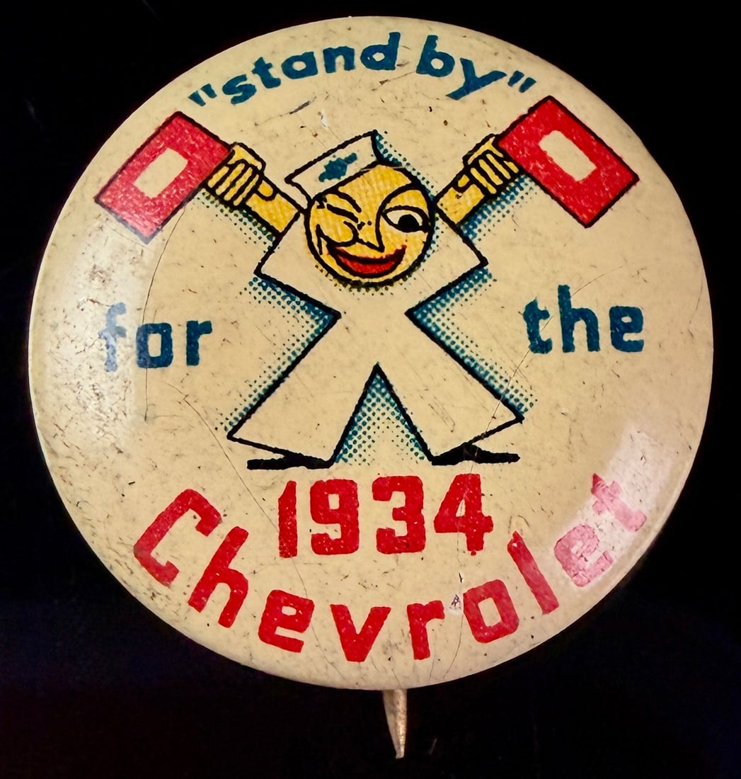 1934 Chevrolet Stand By Advertising Pinback Button by St. Louis Button Co.: This original promotional teaser button was produced for the marketing campaign of the 1934 Chevrolet. The graphic features a winking anthropomorphic mascot in the shape of an X, wearing a sailor cap