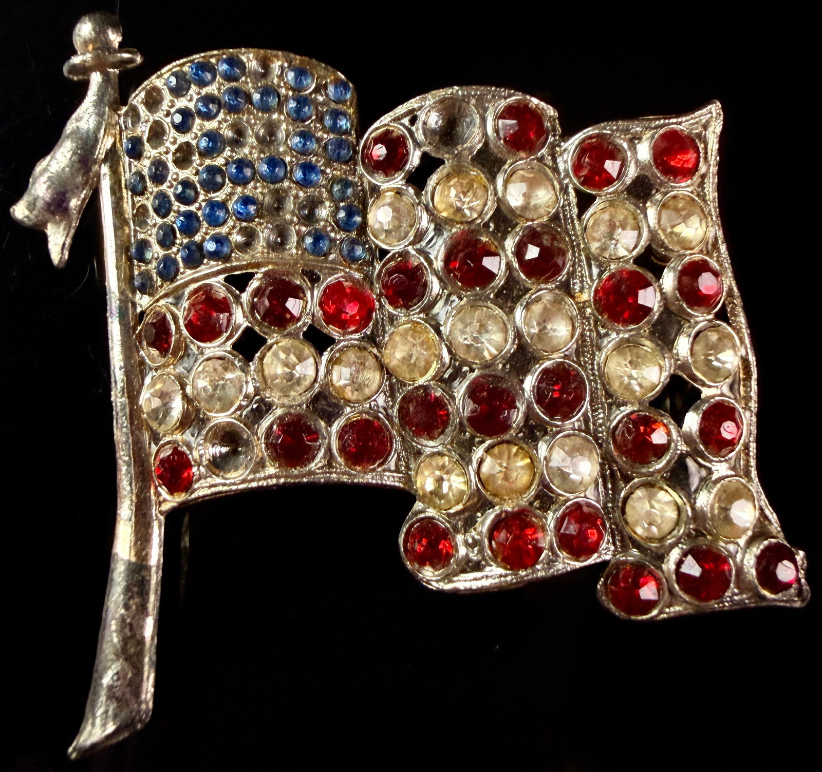 Vintage Sterling Silver and Rhinestone Waving American Flag Brooch: A vintage sterling silver brooch depicting a waving American flag attached to a flagpole. The flagpole features a spherical finial at the top and a decorative draped tassel. The flag is pave-set with 