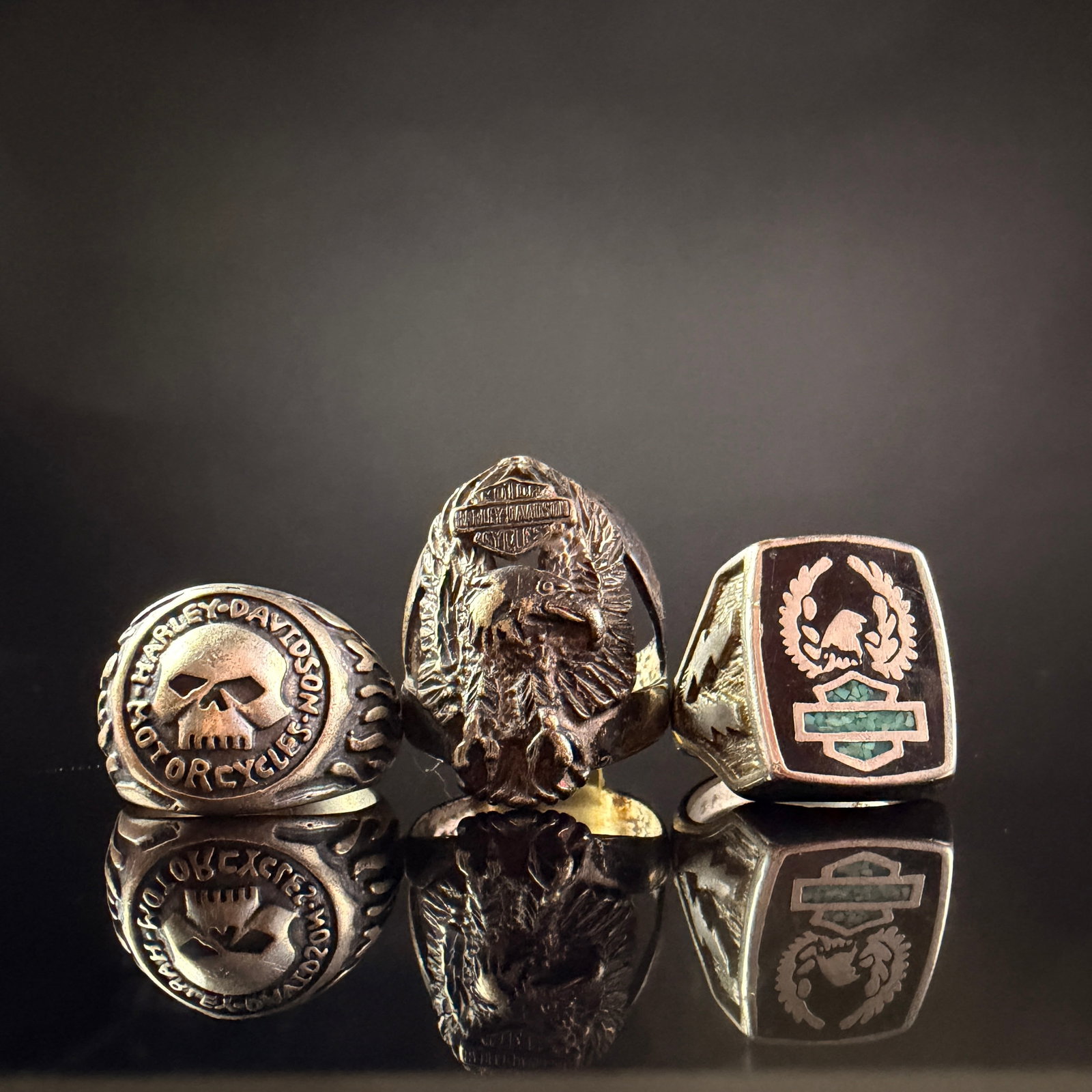 A Trio of Harley-Davidson Sterling Silver Rings: A collection of three sterling silver rings featuring motifs associated with Harley-Davidson. The first ring is a signet style with a central skull design surrounded by a border reading
