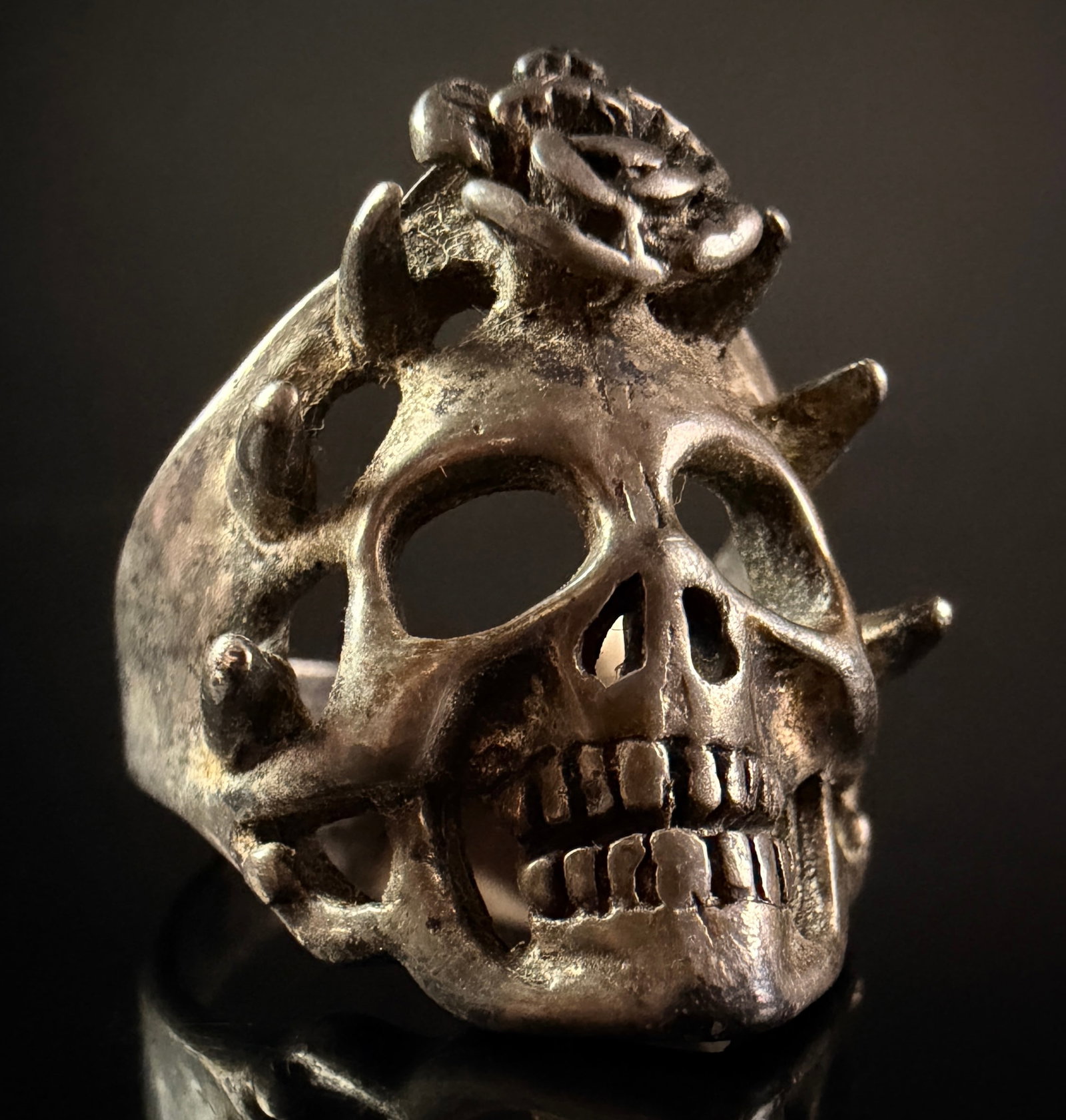 Gothic Style Skull and Rose Biker Ring: A large Gothic-style statement ring featuring a central anatomical skull motif. The cranium is topped with a relief-cast rose blossom and the design is bordered by six prominent curved spikes. The