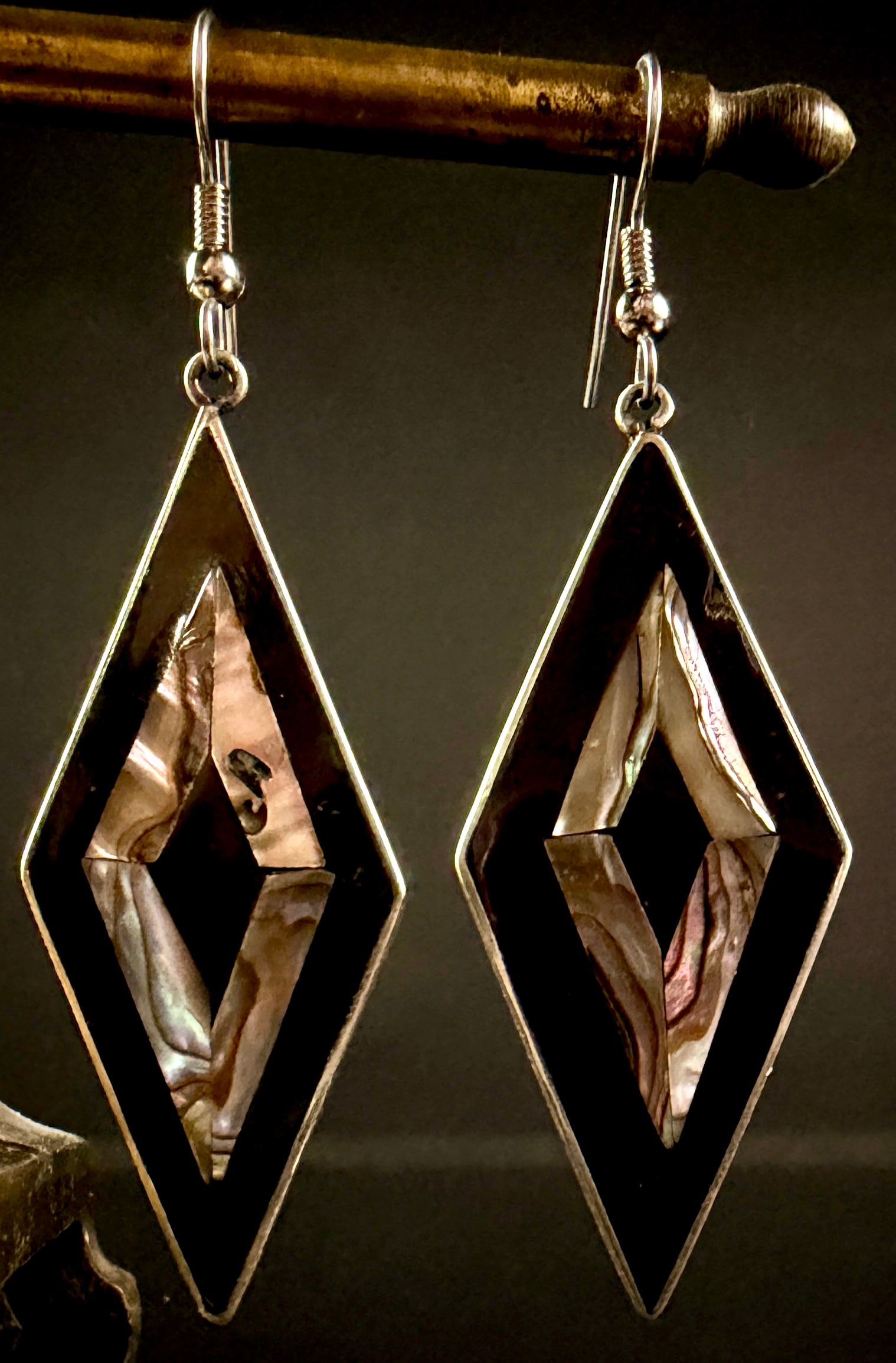 A Pair of Mexican Alpaca and Abalone Shell Geometric Dangle Earrings: A matched pair of large, lozenge-shaped dangle earrings of Mexican origin. Each earring features a diamond-shaped frame with a central diamond cutout, constructed from white metal. The front face is