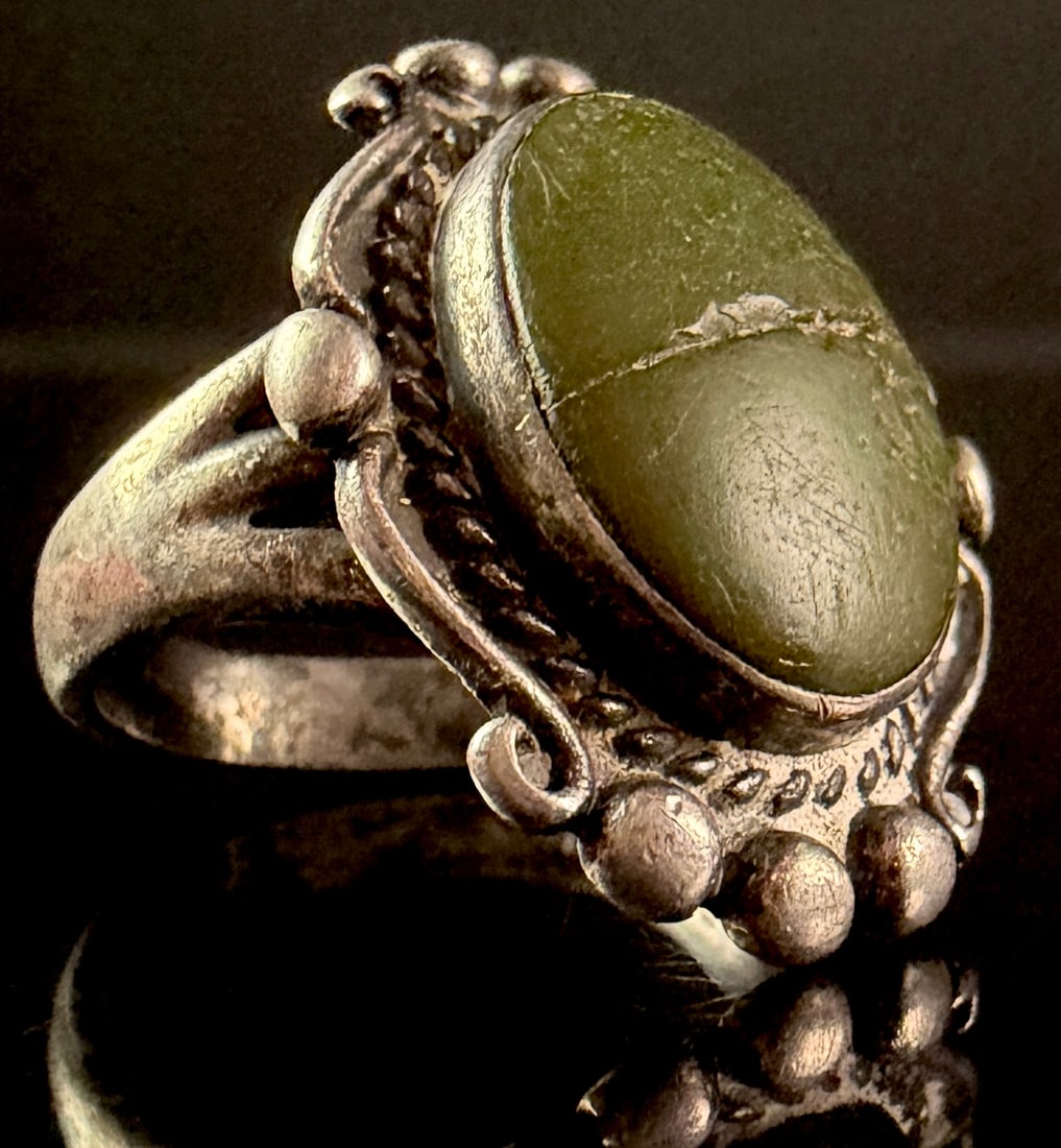 Southwest Style Silver Toned Oval Green Stone Ring: A silver-toned ring featuring a large oval green cabochon stone. The stone is secured in a bezel setting and is surrounded by a decorative rope-twist border and an ornate outer frame composed of