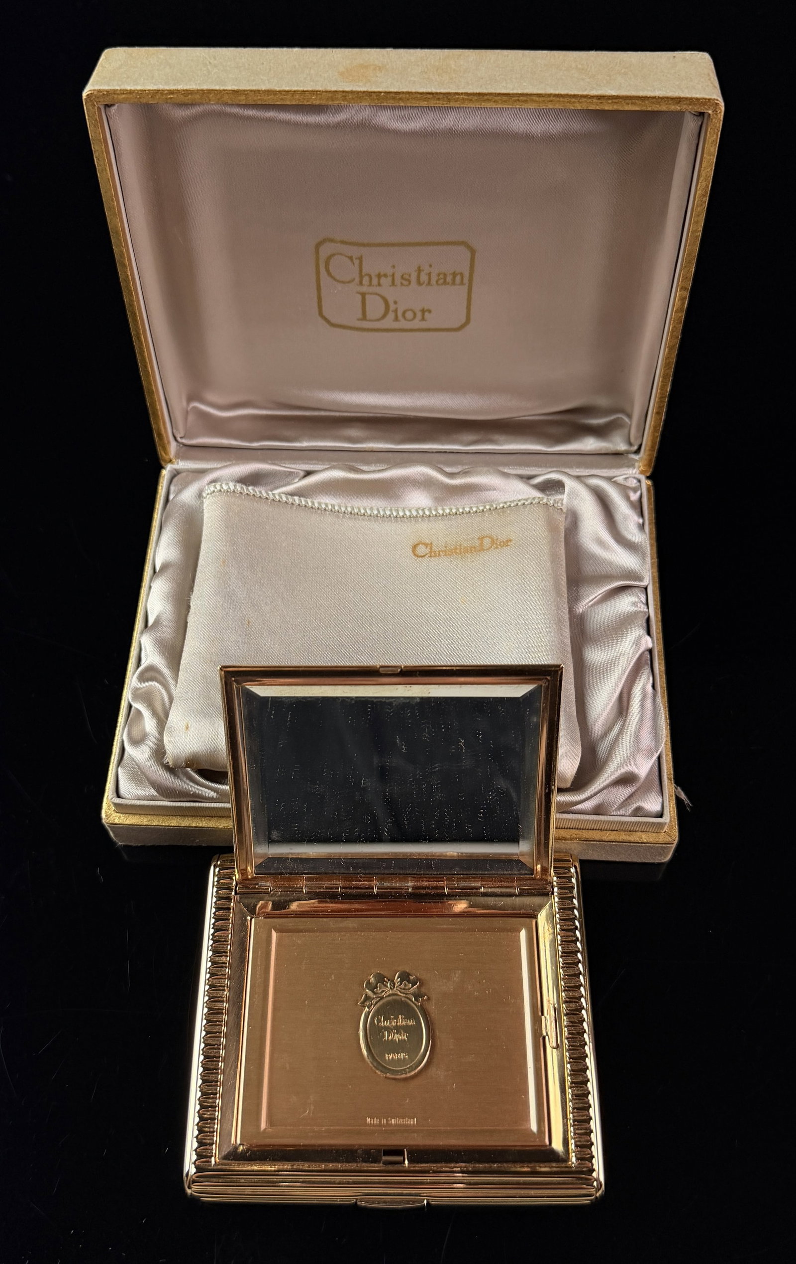 Vintage Christian Dior Gold Tone Ribbed Powder Compact with Original Box and Pouch: A Swiss gold-tone metal powder compact of rectangular form featuring a horizontally ribbed exterior texture. The hinged lid opens to reveal a beveled glass mirror. The interior contains a hinged secon