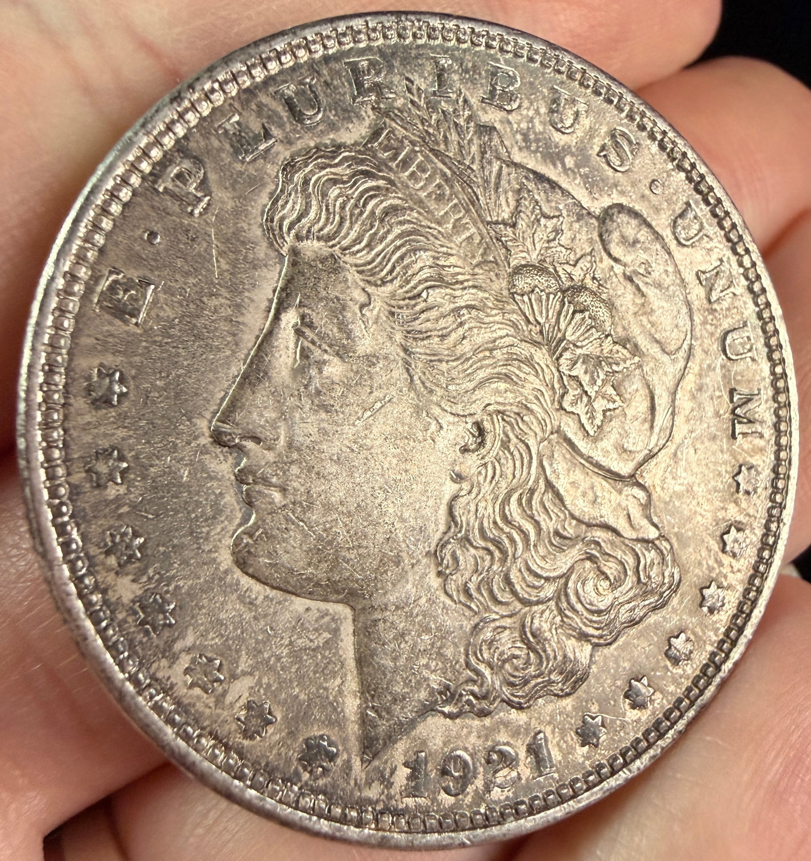 US 1921 Morgan Silver Dollar: A US 1921 Morgan silver dollar composed of 0.900 fine silver. This piece was struck at the Philadelphia Mint, evidenced by the absence of a mint mark on the reverse above the letters DO in DOLLAR. The