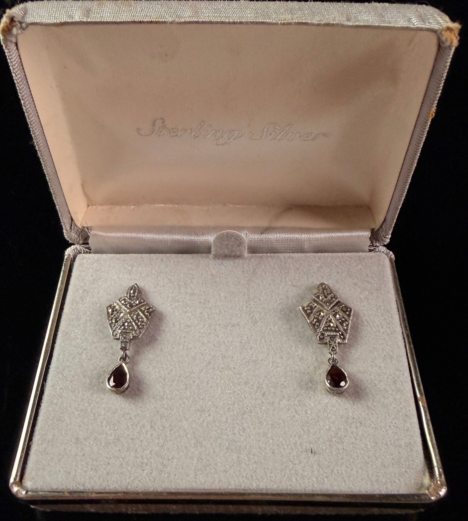 A Pair of Sterling Silver Marcasite and Garnet Art Deco Style Drop Earrings: A matched pair of sterling silver drop earrings featuring an Art Deco inspired design. The upper geometric portion of each earring is shield-shaped and pav?©-set with small faceted marcasite stones. 