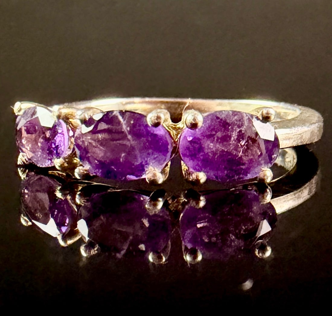 Jessica Lili African Amethyst Sterling Silver Three-Stone Trilogy Ring: A sterling silver three-stone trilogy ring featuring three prong-set, oval-faceted African amethysts. Each gemstone is secured within a four-prong basket setting. The ring is accompanied by a Certific