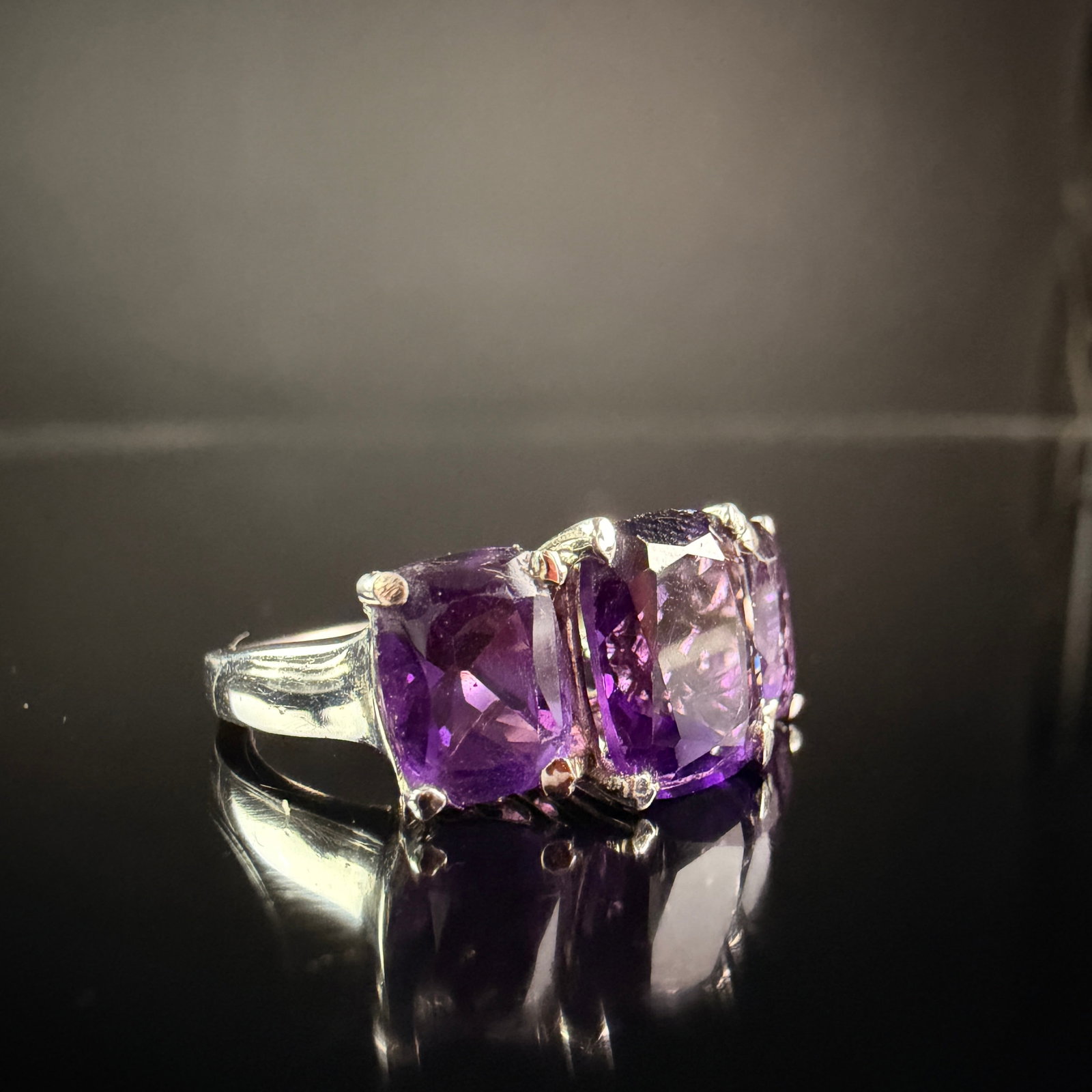 Sterling Silver and Amethyst Three-Stone Cocktail Ring: A sterling silver cocktail ring featuring a trilogy design. The piece is set with three cushion-cut purple amethysts in four-prong settings, with the central gemstone being slightly larger than the tw
