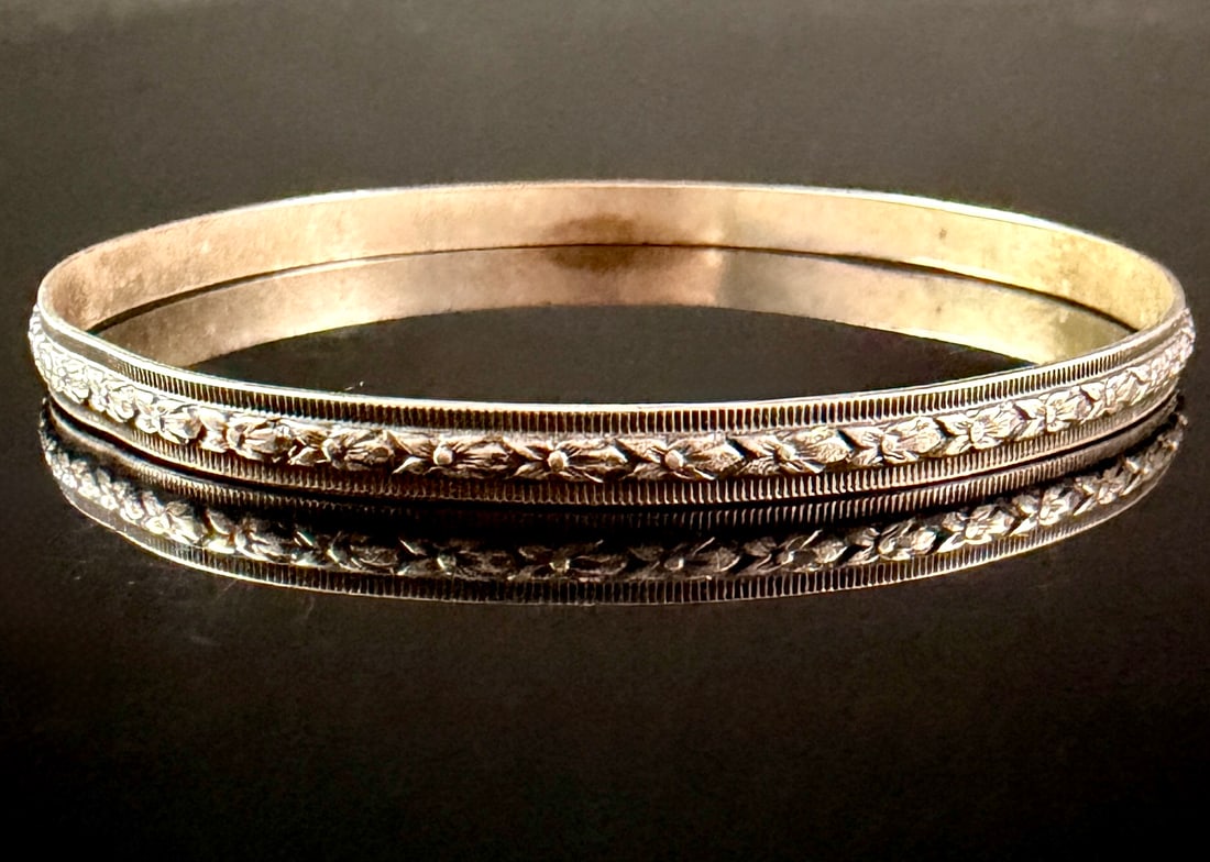 Vintage Sterling Silver Floral Relief Bangle Bracelet: A sterling silver bangle bracelet featuring a solid circular band. The exterior is decorated with a continuous embossed relief pattern of stylized flowers and leaves, set within parallel borders of fi