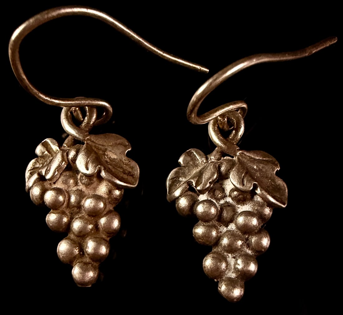 A Pair of Mexican Sterling Silver Figural Grape Cluster Drop Earrings: A matched pair of vintage sterling silver drop earrings, each featuring a three-dimensional cluster of grapes surmounted by two textured vine leaves. The earrings are suspended from simple French hook