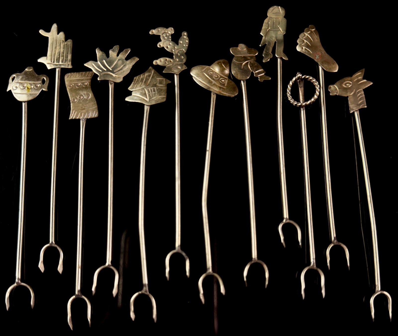 Set of Twelve Taxco Mexico Silver Figural Cocktail Picks: A set of twelve silver cocktail or hors d'oeuvre picks, manufactured in Taxco, Mexico. Each pick features a unique finial representing various Mexican folk art motifs, including a teapot, organ pipe c