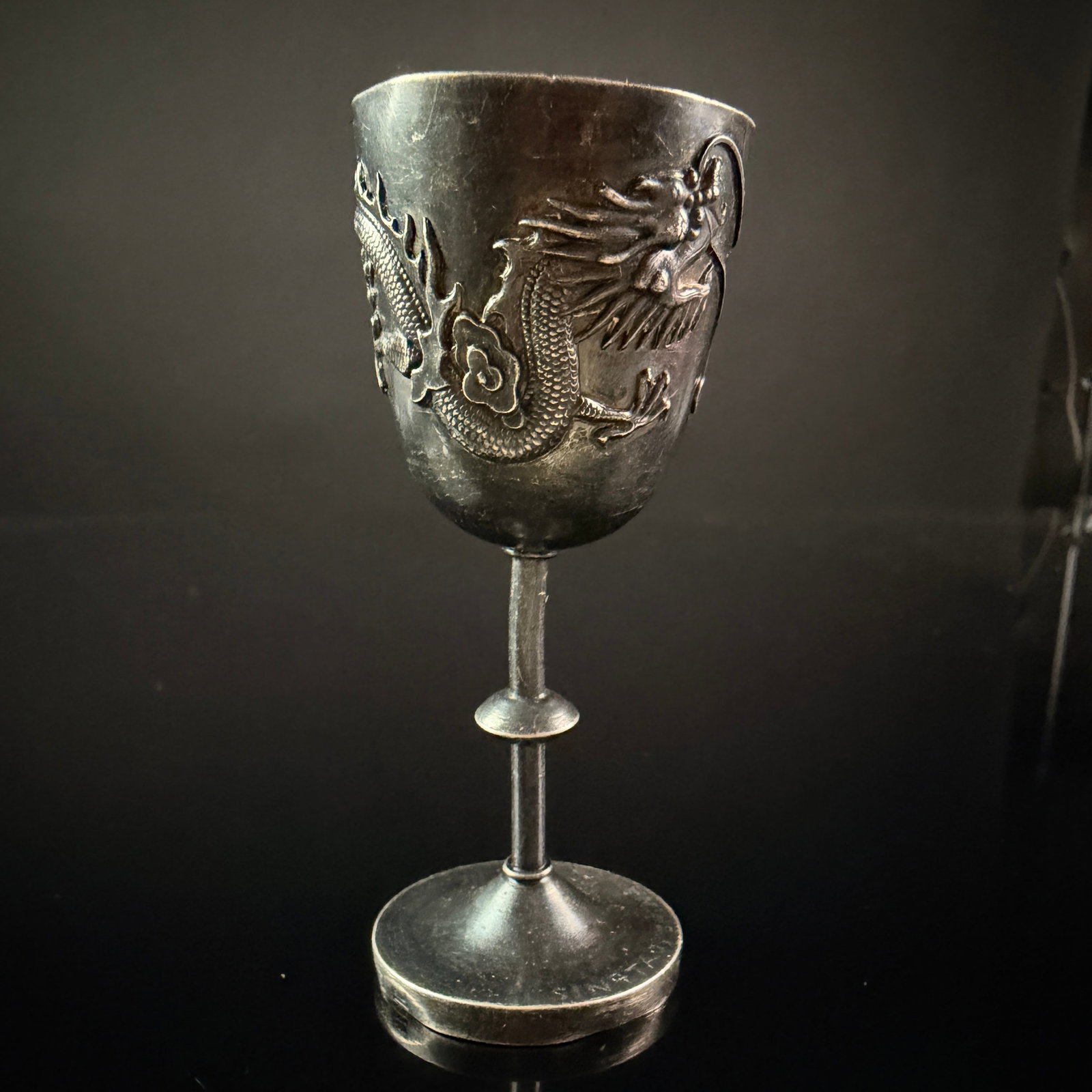 Chinese Export Silver Dragon Cordial Cup, Tsingtau, China, 1945: A Chinese export silver cordial cup featuring a bowl with high-relief repousse decoration of a four-clawed dragon chasing a flaming pearl among stylized clouds. The cup is supported by a slender stem 