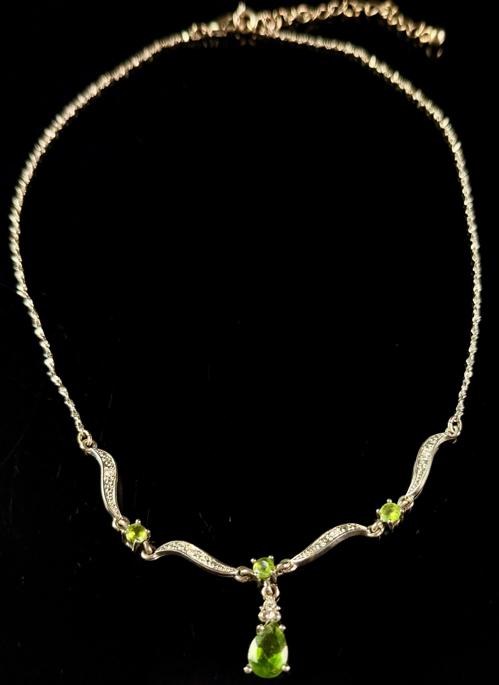 Sterling Silver and Peridot Gemstone Y-Necklace: A sterling silver necklace featuring four faceted light green peridots. The design consists of a central pear-cut peridot drop and three round-cut peridots integrated into decorative S-curved links. T
