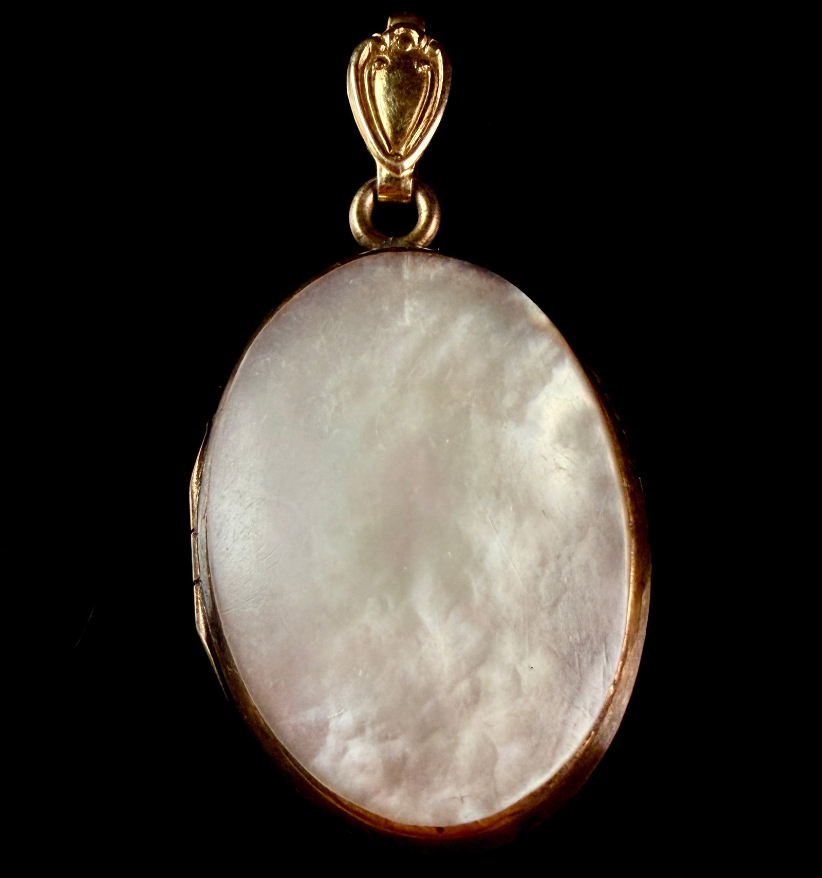Twelve Karat Gold-Filled and Mother-of-Pearl Oval Locket Pendant: This oval locket pendant is constructed from 12 karat gold-filled metal and features a polished mother-of-pearl inlay on the front face. The mother-of-pearl exhibits natural white and silver iridescen
