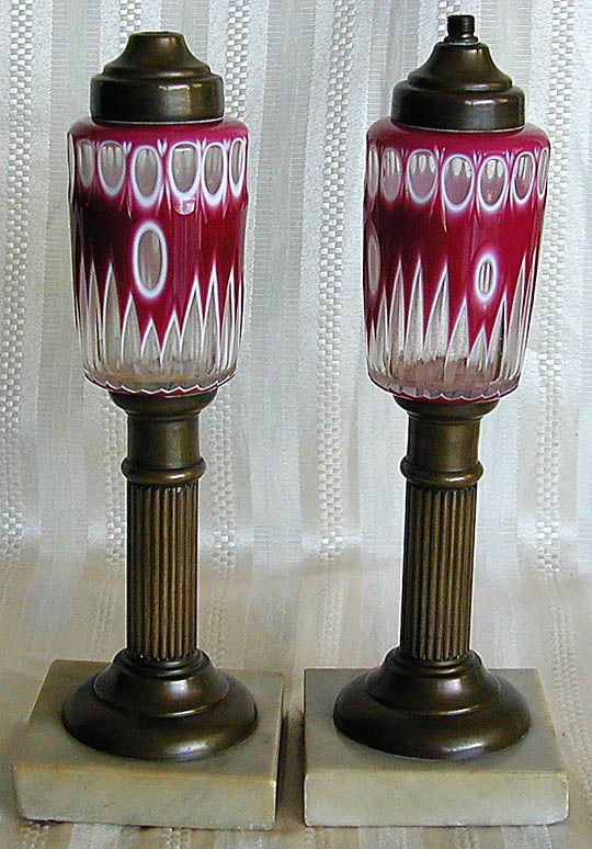 PR ANTIQUE CASED RUBY CUT TO CLEAR OIL LAMPS: A pair of Antique cased ruby cut to clear glass oil lamps mounted on fluted bronze column bases with oil receptables in a deep cut glass design and mounted on square marble bases, good condition, 10.2